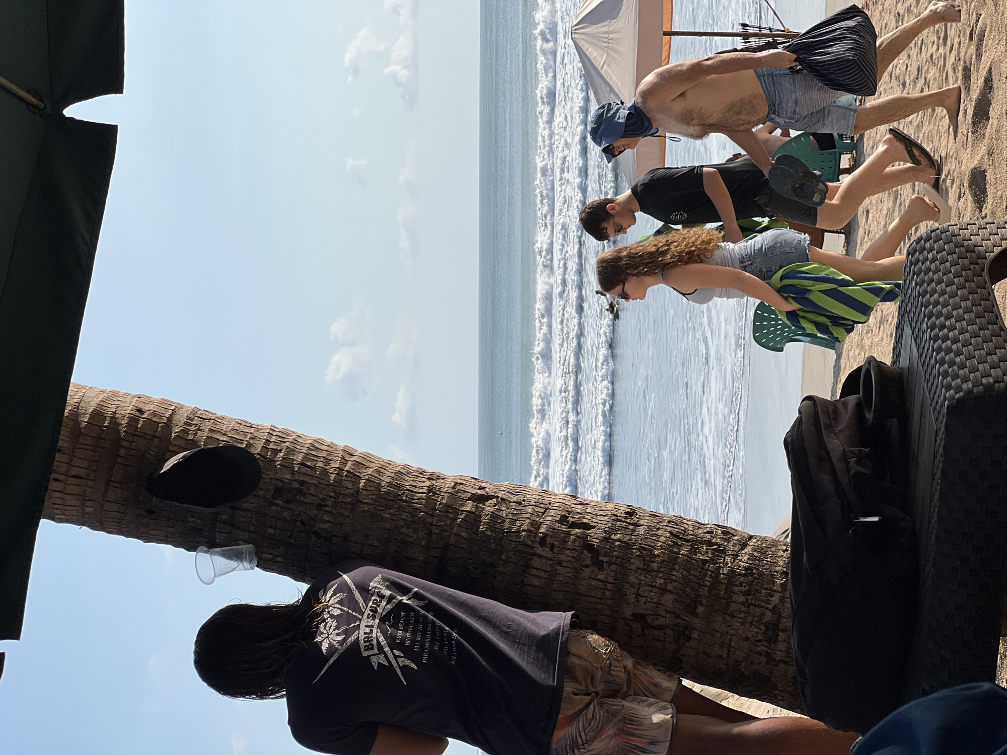 The image depicts a sunny day on the beach in Kuta, Bali, Indonesia, with the Indian Ocean in the background. The foreground shows a portion of a palm tree trunk with a black hat and a plastic cup attached to it. To the left, a person with long, dark hair, wearing a dark t-shirt with a "Bali Surf" graphic and patterned shorts, is partially visible, leaning against the tree.
In the mid-ground, three people are walking along the sandy beach towards the right. The first person is a woman with long, curly brown hair, wearing a light blue tank top and denim shorts, carrying a striped towel. Next to her is a man wearing a black t-shirt and dark shorts, holding flip-flops. The third person, a man, is wearing a blue sun hat and striped blue shorts, carrying a striped bag. They are walking on the sand, with the ocean waves in the background.
The environment is a beach with fine sand. The ocean is a vibrant blue with white-capped waves. The sky is clear blue with scattered white clouds. In the far background, there are some distant figures in the water and on the beach, suggesting other beachgoers. There is also a beige beach umbrella visible on the right side of the image.