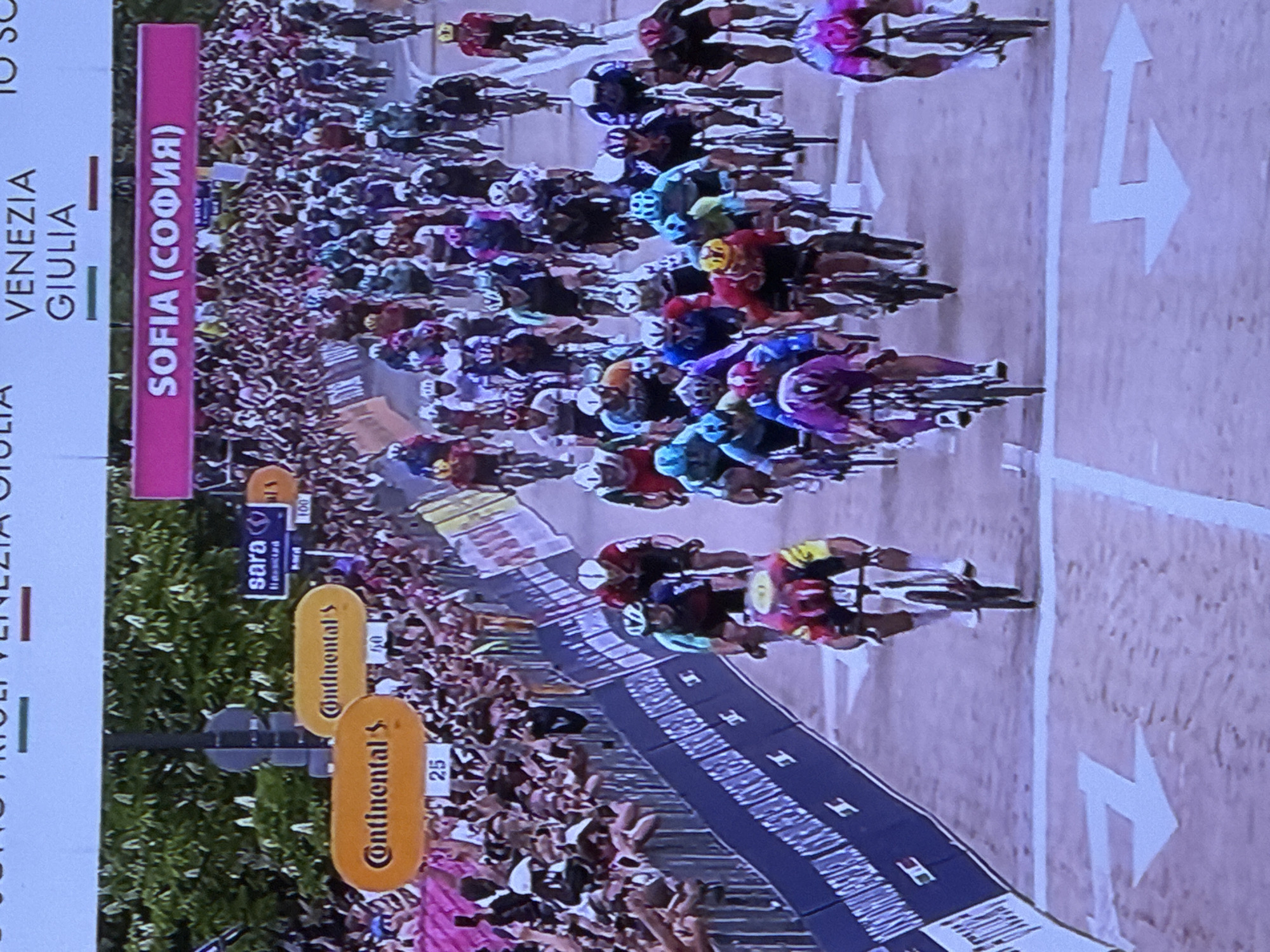 The image shows a large group of cyclists racing on a road. They are closely packed together, many wearing helmets in different colors. The sides of the road are lined with spectators. Signs labeled "Continental's" and "sara" are visible. The road has white arrows painted on it. A sign displays the text "VENEZIA GIULIA" above "SOFIA (COФИЯ)", indicating this segment of the race is in Sofia, Bulgaria.