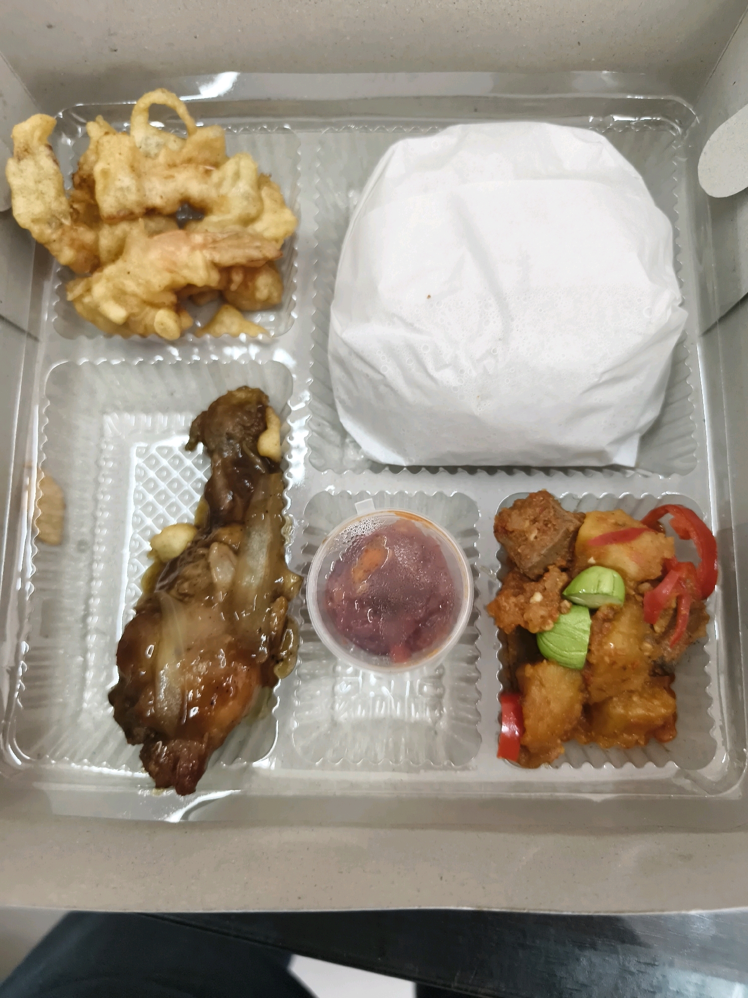 A top-down, close-up, eye-level shot captures a takeout meal box. The box is divided into four compartments, with each section containing a different food item. On the top left, a pile of golden-brown, fried shrimp rests. To its right, a white paper-wrapped mound of rice dominates the compartment. The bottom left section holds a piece of glazed, dark meat, possibly chicken or duck, with what appear to be garlic cloves. To the right of that, a small, clear plastic container filled with a reddish sauce sits. The final compartment on the bottom right contains a stir-fry mixture of cubed meat, potatoes, slices of red chili, and green beans. The meal box is presented against a neutral, light-colored background, suggesting it was photographed in an indoor setting.