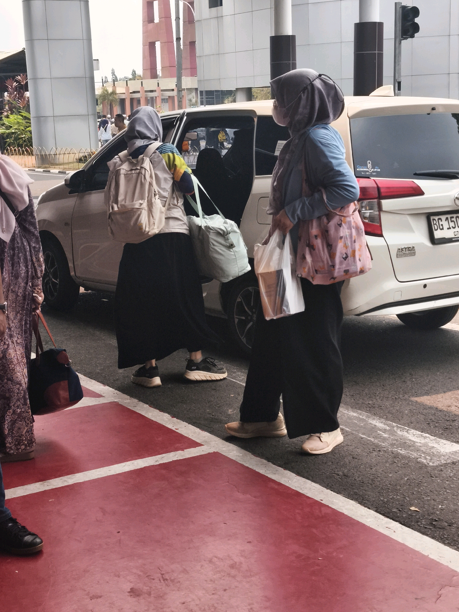 The image captures a street scene in Palembang, Indonesia, likely near a transportation hub given the presence of a white car with its door open. Two women, dressed in hijabs and modest clothing, are the primary subjects. The woman on the left is wearing a patterned purple dress and carrying a red, blue, and black duffel bag. She is standing near the rear of the car. The woman on the right is wearing a blue jacket over a patterned pink top and black pants. She has a gray hijab and is carrying a clear plastic bag with items inside and a pink patterned tote bag. She is standing closer to the open car door. The car appears to be a Toyota, with a license plate starting with "BG 150". In the background, a tall, angular building with red accents dominates the skyline, suggesting an urban setting. The ground is paved with asphalt and marked with red and white lines, indicating a pedestrian crossing or walkway. The overall atmosphere suggests a moment of transition, possibly arriving at or departing from a destination.