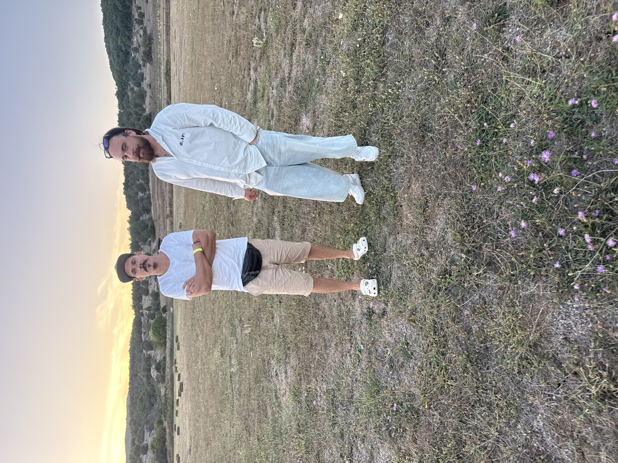 Two men are standing in a field with dry grass under a pale sunset sky. The man on the left is wearing a white t-shirt, beige shorts, a black fanny pack, and white Crocs. He has his arms crossed and is wearing a baseball cap and has a mustache. The man on the right is wearing a light-colored striped button-down shirt, light-colored wide-leg jeans, and white sneakers. He has sunglasses perched on his head and a beard. In the background, there are rolling hills covered in trees and some hay bales. The ground is covered in dry grass and small purple flowers. The scene appears to be in a rural or natural setting.