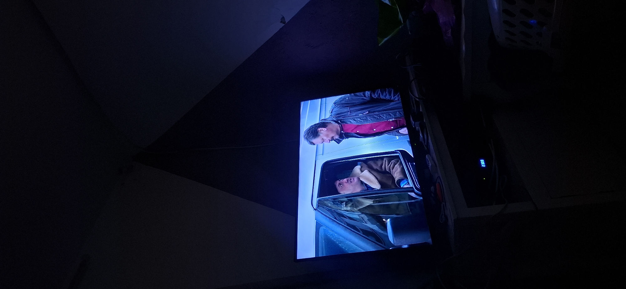The image shows a television screen displaying a scene with two men. The man on the left is inside a white van, wearing a brown jacket with a fur collar. He is looking towards the man on the right. The man on the right is standing outside the van, wearing a red shirt and a dark jacket, and is looking at the man in the van. The television is placed on a shelf in a dark room. The room appears to be a bedroom, with dark walls and a ceiling that forms a corner above the television. There is some electronic equipment and other items on the shelf below the television. The time "23:08" is visible on a digital display below the television. The image is captured from a slightly upward angle, suggesting the viewer is looking at the television from a seated or lying position.