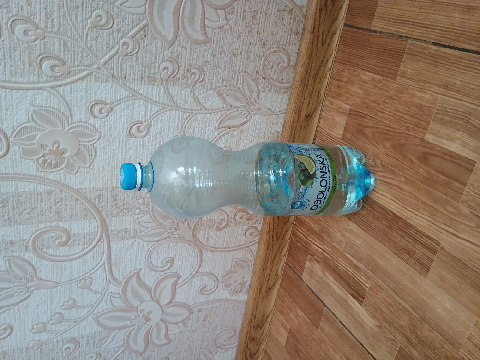 A plastic bottle of "Obolonska" brand sparkling water lies horizontally on a wooden floor next to a wall with floral wallpaper. The bottle is mostly clear with some condensation, and the water inside is a light blue color. The label on the bottle is partially visible and reads "Obolonska" in large letters, with smaller text beneath that includes "sparkling non-alcoholic drink" and "Lime And Mint." There's also a small circular logo with "kcal" inside. The wallpaper has a textured, embossed floral pattern in light cream and beige tones. The floor appears to be made of wood planks laid in a herringbone pattern, with some dark lines between them suggesting grout or seams. A portion of a wooden trim piece is visible to the right of the bottle. The lighting suggests it is daytime, and the indoor setting appears to be a home or possibly an office space. No people are visible in the image, and the overall scene is static. The location is specified as Zaporizhzhia, Ukraine.