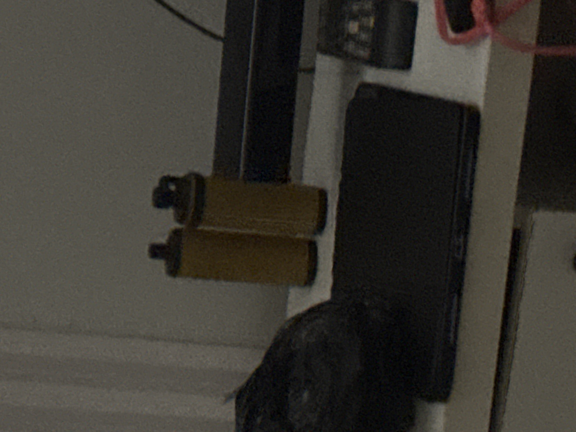 A close-up, slightly grainy, eye-level shot focuses on a desk with various objects. In the foreground, a black laptop is open, its screen facing away from the viewer. To the right of the laptop, a black object with small buttons, possibly a remote or calculator, is visible. Further to the right, a bright pink cord loops and is partially visible. Behind the laptop and to the left, a black object, possibly a bag, is present. On the desk, standing upright, are two cylindrical objects with yellow-brown coloring, resembling small canisters. The background consists of a textured, pale surface, possibly a wall or curtain. The scene is indoors, with soft, diffused lighting. The location is specified as Abuja, Nigeria.