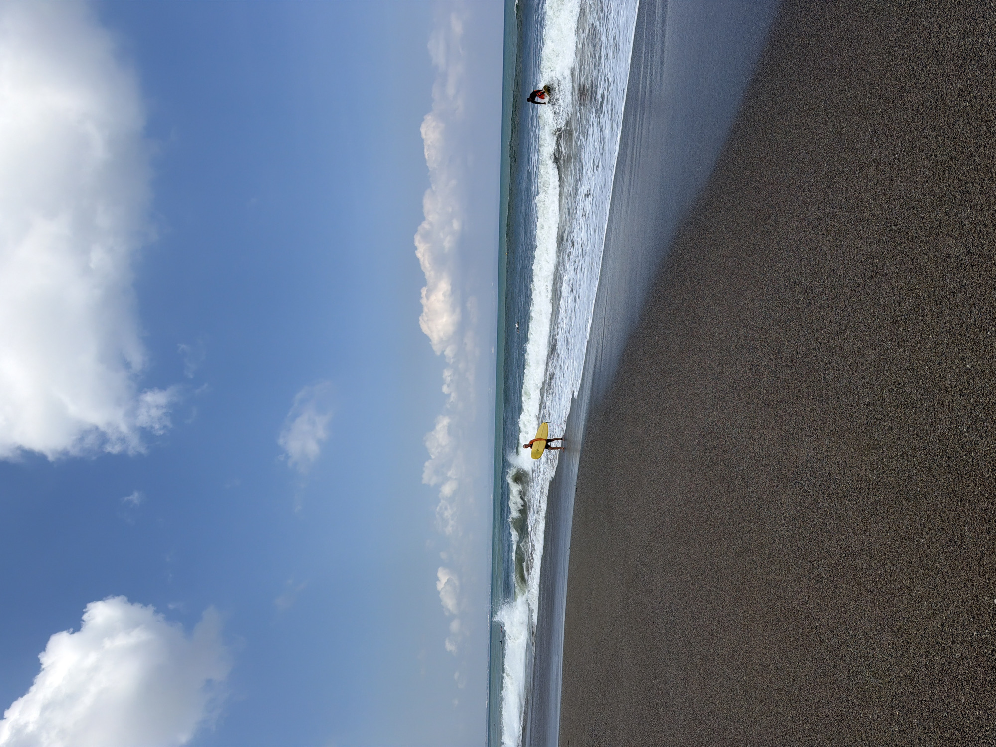 On a sunny day in North Kuta, Indonesia, two individuals are engaged in watersports on a black sand beach. The tide is coming in, with moderate waves breaking.
In the foreground, a person in a red shirt and black shorts walks along the wet sand, carrying a yellow surfboard. Further out in the water, a second person is visible, also with a surfboard, riding a wave. The ocean is a deep blue-green, and the sky is clear with scattered white clouds. The beach itself is composed of dark, fine-grained sand. The horizon line is visible in the distance, with a hint of land on the far left.