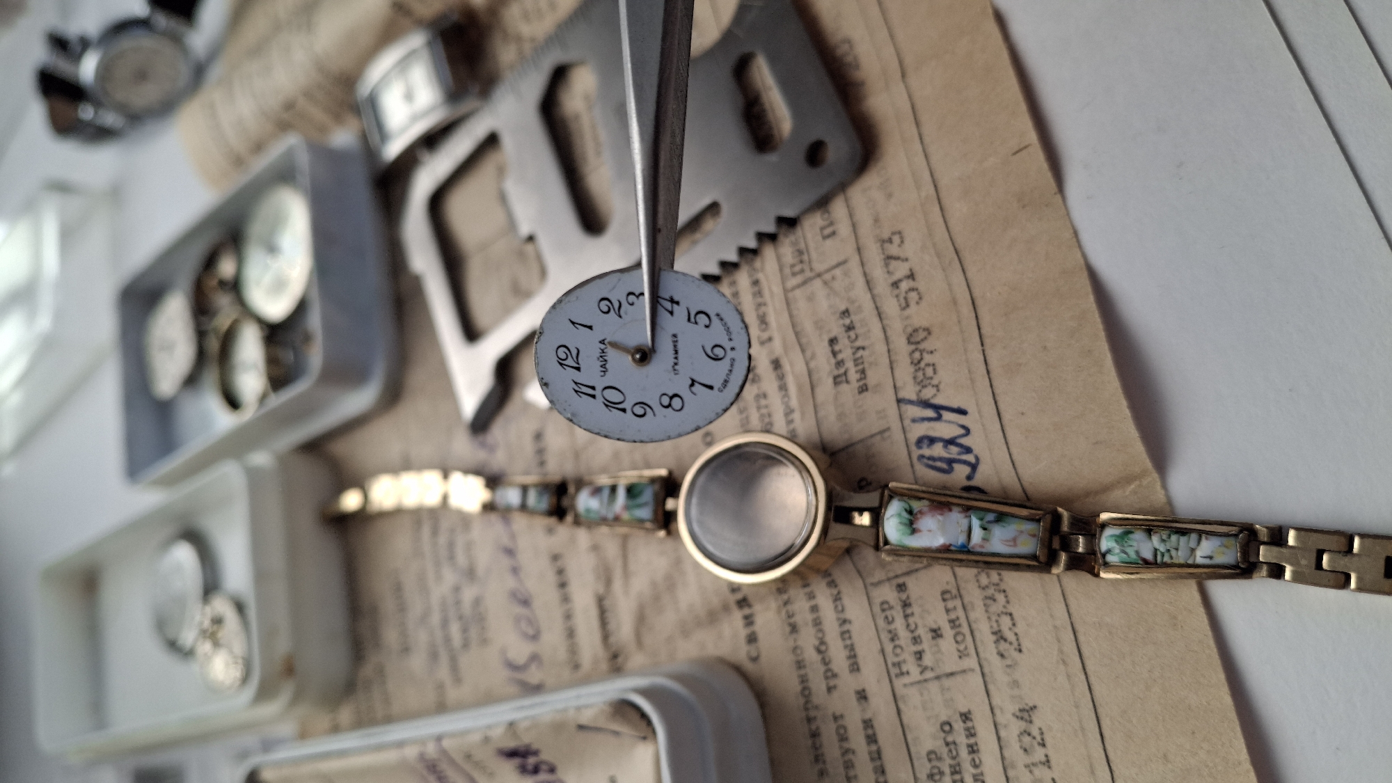 A pair of metal tweezers holds a small, circular, white watch dial in the foreground, centered on the right. The dial displays black Arabic numerals 1 through 12, with a black hour hand positioned between 4 and 5, and a black minute hand pointing directly at 12. Cyrillic text on the dial reads "ЧАЙКА" (Chaika), "17 КАМНЕЙ" (17 Jewels), and "СДЕЛАНО В РОССИИ" (Made in Russia).
Immediately to the left of the tweezers is a gold-colored ladies' wrist watch with an ornate bracelet. The bracelet consists of rectangular segments, some of which feature multicolored (white, green, blue, and reddish-pink) abstract or floral designs. The watch case is round and gold-colored, with a slightly domed, translucent, possibly frosted crystal that obscures the internal mechanism.
All items rest on an aged, creased, light brown paper document featuring black printed Cyrillic text and handwritten black numbers. Visible phrases include "Дата выпуска" (Date of issue) and "Номер участка и контр." (Section and control number). Handwritten numbers "924 / 0890 5173" are prominent in the lower right portion of the document.
In the blurred background, to the left, is a white rectangular plastic container holding several small, round, metallic watch components. To the right, a grey rectangular plastic container also holds multiple small, round, metallic objects. A silver-colored metal multi-tool with various cut-outs and a serrated edge is partially visible to the right of the central watch dial. Further back, two additional watches are out of focus: a rectangular silver-colored watch and a round watch with a black strap.
The scene depicts a watch repair or collection setup. The presence of Cyrillic text indicating "Made in Russia" confirms the country of origin or context. The specific city of Kempelevo, Russia, as mentioned in the prompt, cannot be verified or identified from the visual content of the image.