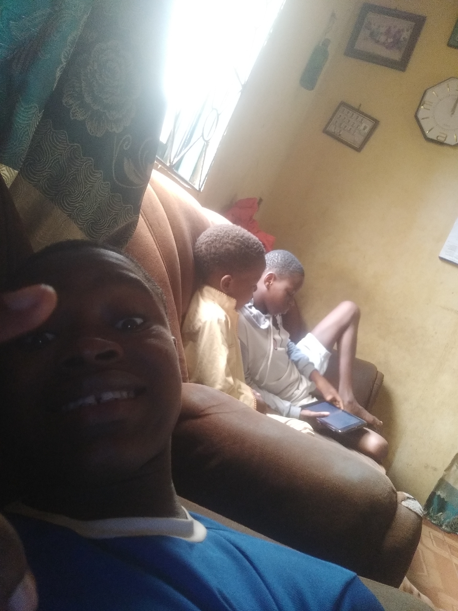 Three young boys are seated on a brown sofa in a room. The boy in the foreground wears a blue shirt and is taking a selfie. The two boys behind him are using a tablet. The wall behind the sofa is light yellow and adorned with framed pictures and a clock. A curtain is visible on the left side of the image. The location is Unknown, Unknown.