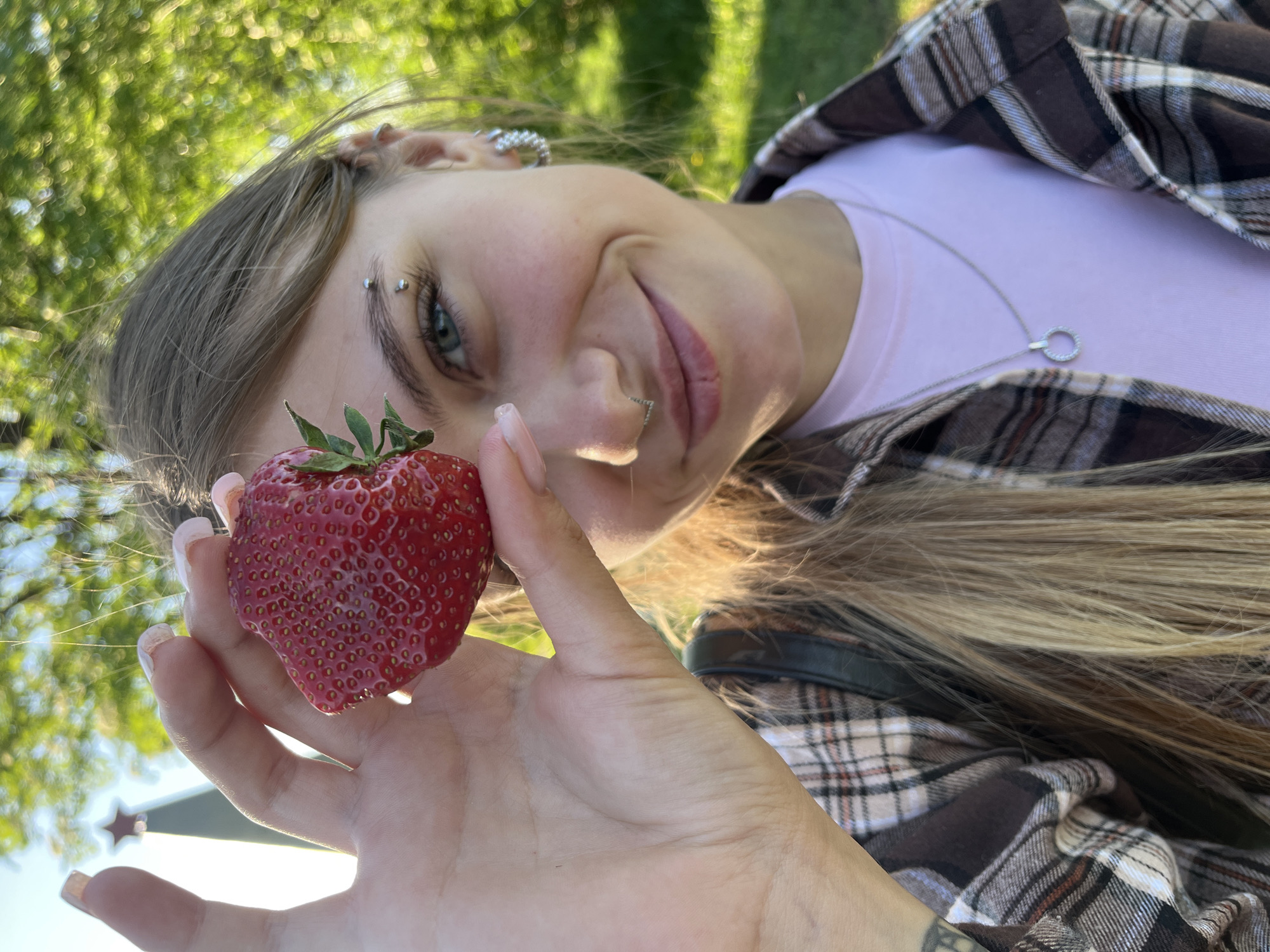 A young woman is holding a strawberry in her right hand. She has blond hair and is wearing a pink shirt and a brown and white plaid jacket. She also has multiple piercings in her ear, eyebrow, and lip, and a silver necklace. The background is blurred, but appears to be a park or outdoor area with green trees and possibly a building with a star visible. There is nothing in the photo to indicate the city of Фоки, Russia.
