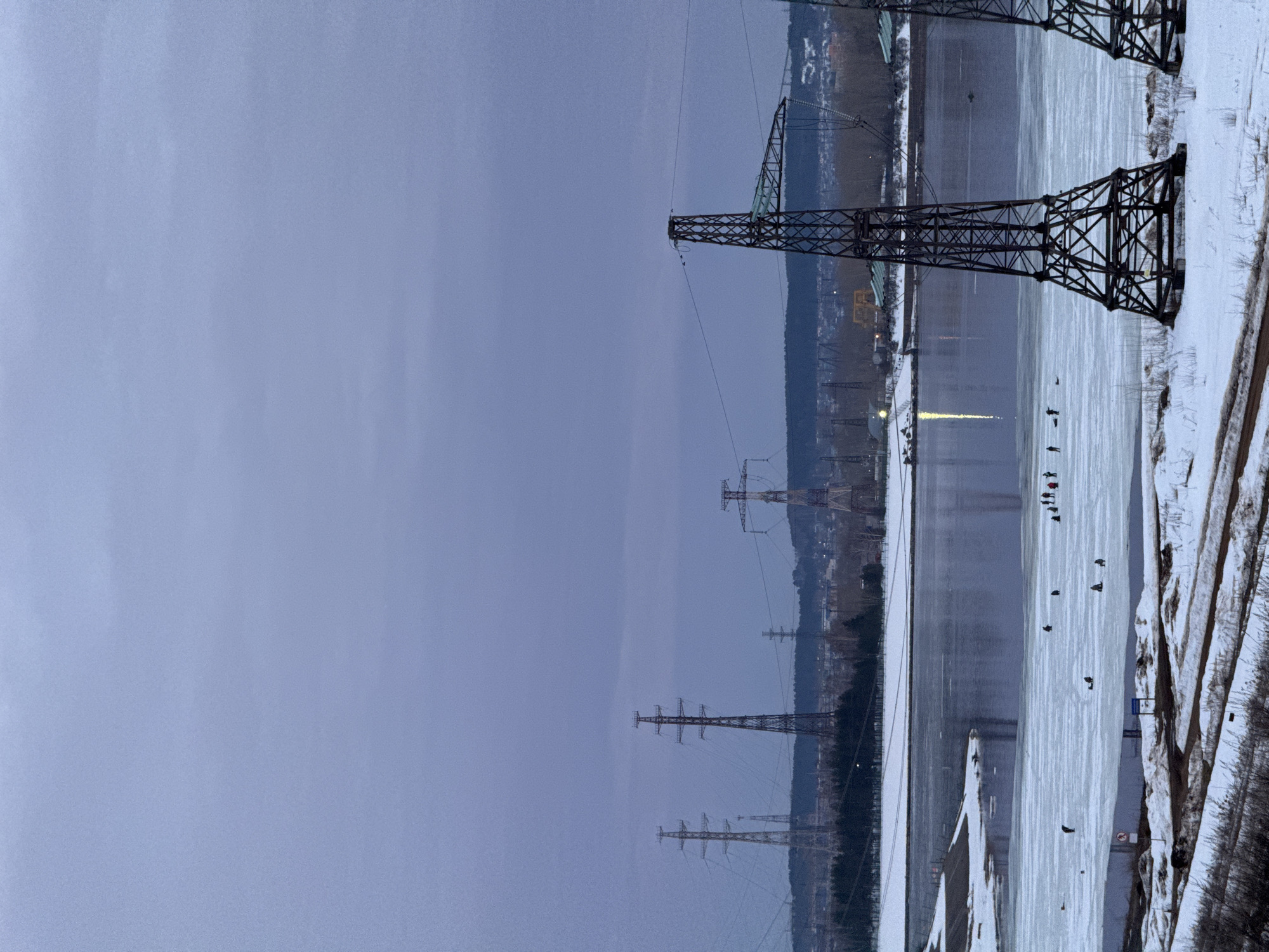 The image depicts a winter or early spring scene in Chaikovsky, Russia, overlooking a partially frozen body of water, likely the Votkinsk Reservoir or Kama River. The sky is uniformly overcast, exhibiting a pale blue-grey hue.
In the foreground, snow-covered ground slopes towards the water's edge, revealing patches of dry vegetation and dirt paths. A red circular "no entry" sign with a diagonal bar and a smaller blue informational sign are visible on the left side near the shore. Two large, dark brown lattice steel transmission towers (pylons) stand prominently on the right, extending from the bottom of the frame and supporting power lines.
The mid-ground features a wide expanse of water, significantly covered by irregular sheets of white ice interspersed with areas of dark, open water. Approximately 10 to 15 individuals are visible across the ice surface, engaged in ice fishing. They are dispersed, mostly standing or kneeling, near what appear to be fishing holes. Reflections of distant lights are visible on the open water towards the right.
The far bank of the waterway is characterized by a mix of snow-dusted terrain, dark evergreen trees, and bare deciduous trees. Beyond these, the urban and industrial infrastructure of Chaikovsky is discernible, including buildings and lights. Several additional high-voltage transmission towers of various designs are positioned across the distant landscape, with some appearing to span the water further along the horizon.