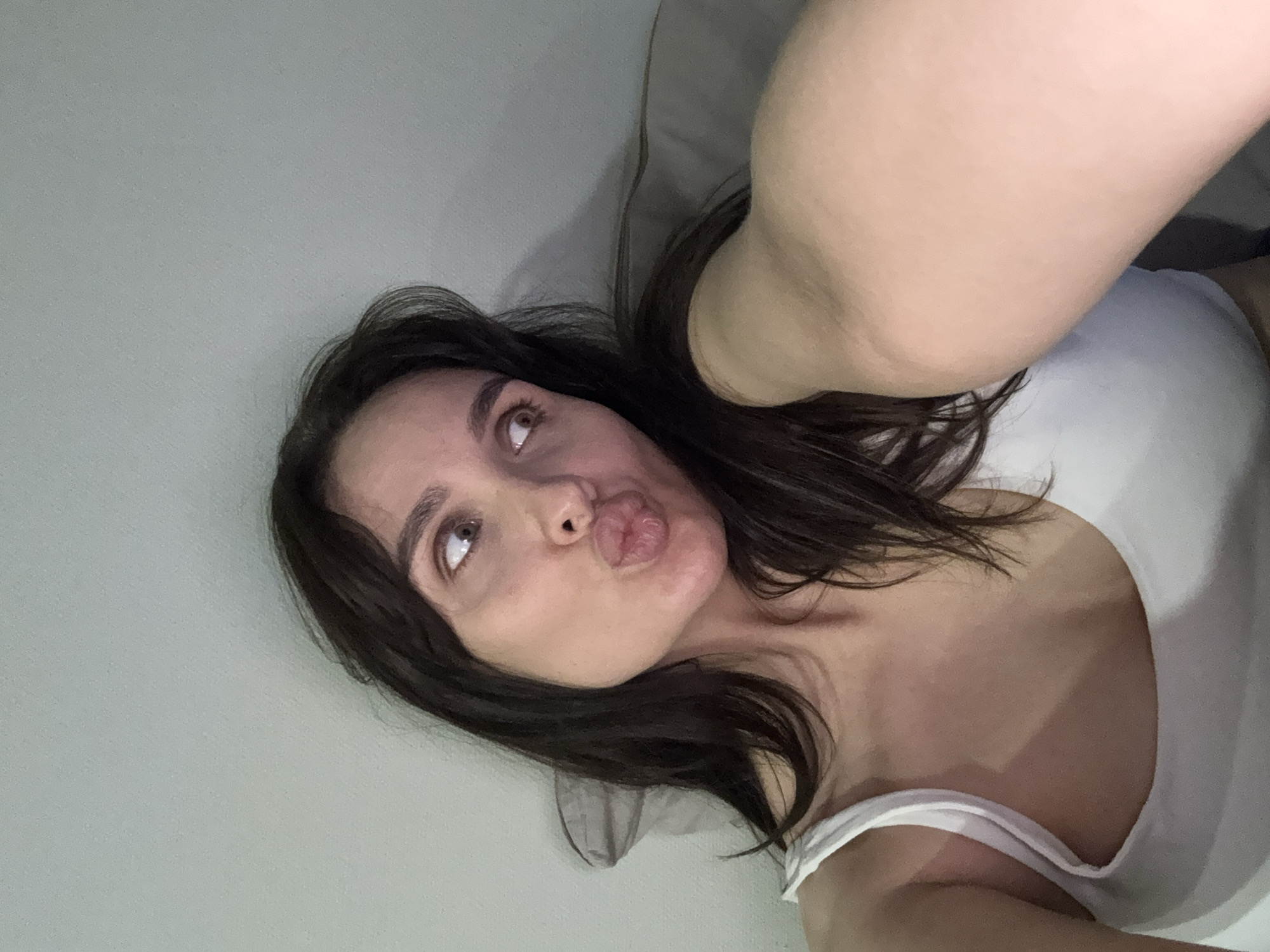 A woman with dark hair lies on a grey surface, possibly a bed or sofa, and wears a white tank top. She looks upwards and slightly to the left with a pouting expression. The background is a plain light grey wall. No information is present to definitively place the scene in the city of Chaikovsky, Russia.