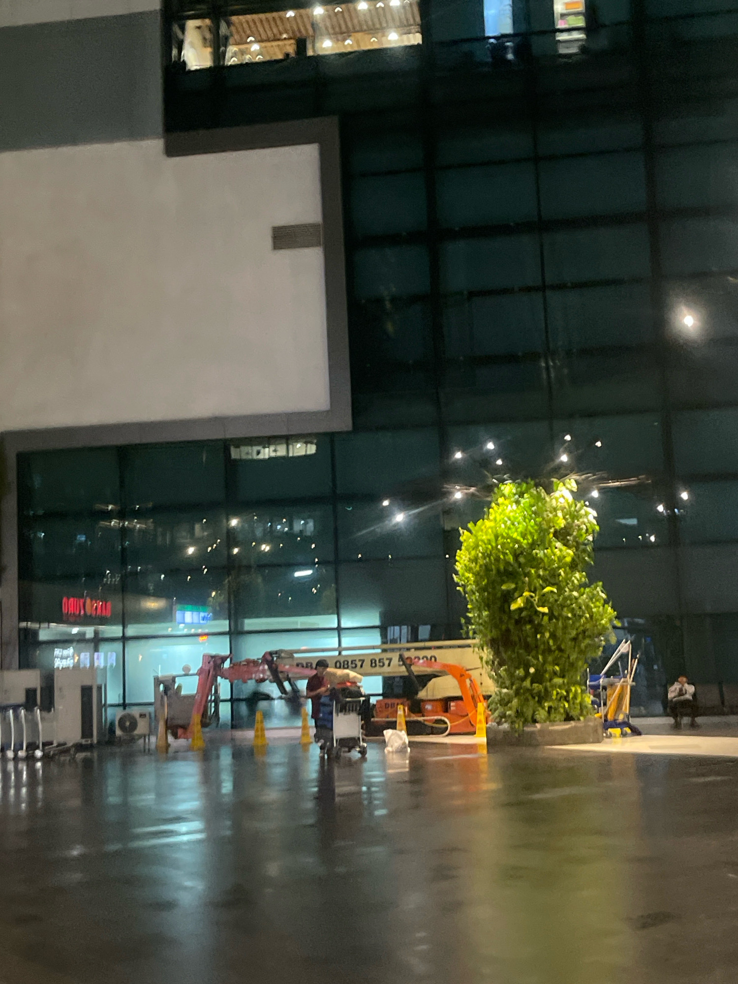 The image depicts an outdoor scene at night, possibly a public area or entrance of a building. The ground is wet, reflecting the lights from the surroundings, suggesting it has recently rained or is currently raining. In the foreground, there are several shopping trolleys and a person pushing a trolley. A piece of construction equipment, possibly a cherry picker, is visible. There are also traffic cones and an outdoor air conditioning unit. In the background, a large building with reflective glass windows is present, showing interior lights and possibly reflections of the exterior. A large green plant is situated in the midground. A person is seen sitting on a bench on the right side of the image. The overall lighting suggests it is nighttime. The image is of low quality and blurry, making precise identification of details difficult. There is no visual information in the image that specifically places it in Pajang, Indonesia.