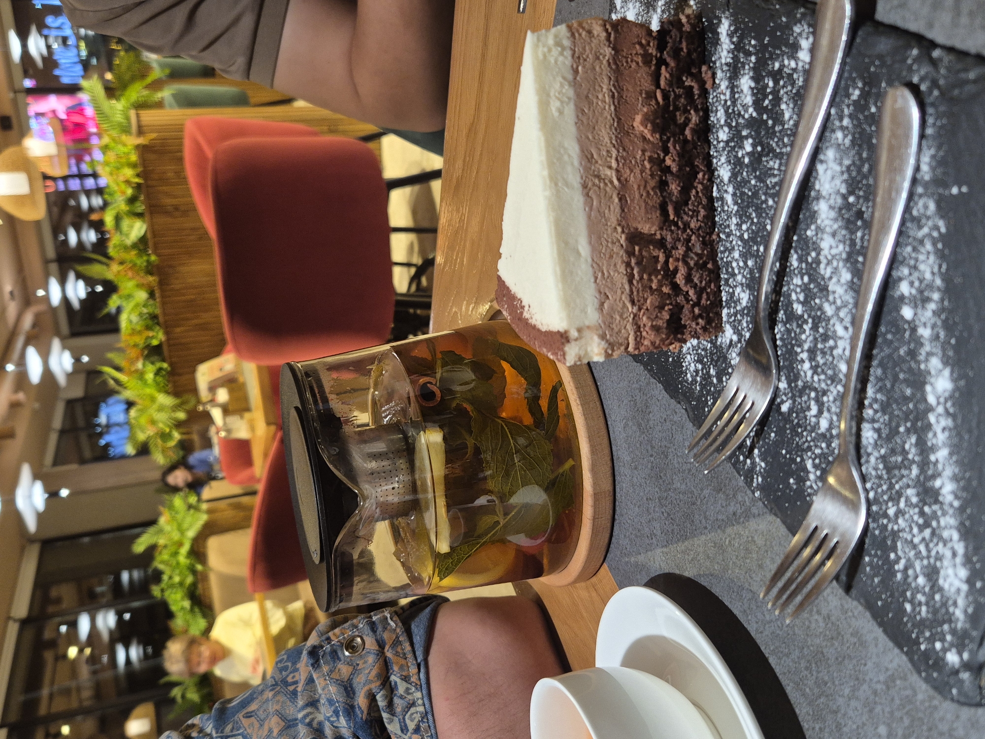 The image depicts a table setting at a cafe, likely in Kokshetau, Kazakhstan. A slice of three-layered cake (chocolate, cream, chocolate) sits on a dark slate plate dusted with powdered sugar, accompanied by two silver forks. Next to the cake is a glass teapot filled with tea, including mint leaves and lemon slices. There's a stack of white ceramic dishes (possibly a cup and saucer) as well. A person wearing a patterned denim shirt is partially visible, as is another individual wearing a brown top. In the background, one can discern red chairs and plants that are part of the cafe's decor. Through the cafe window, city lights and other buildings are visible.