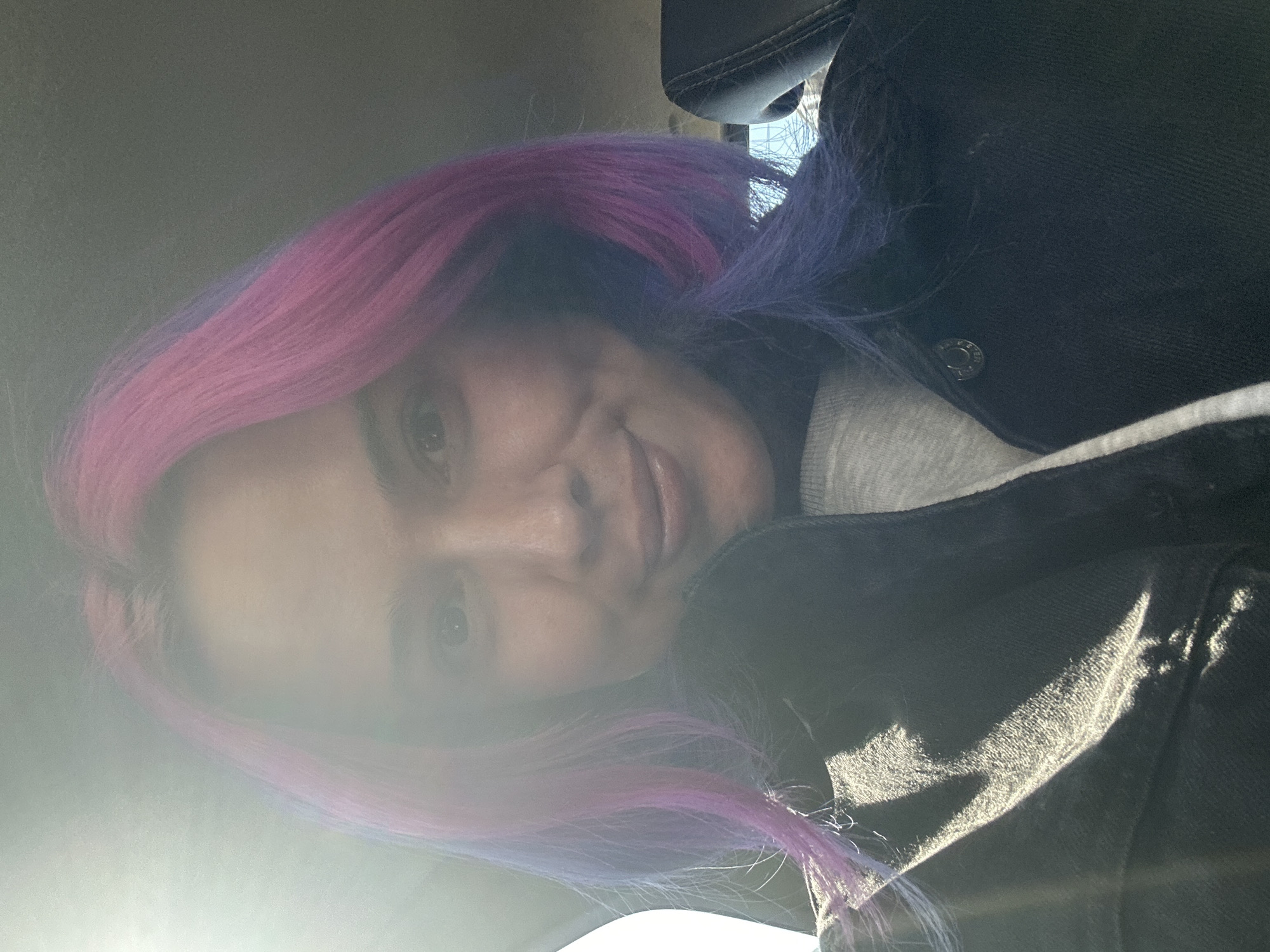 The image presents a close-up, sideways-oriented portrait of a person, likely taken as a selfie, situated within what appears to be a car interior. The subject has distinctively colored hair, with vibrant pink or magenta visible on one side and purple or blue on the other. A slight, gentle smile is present on their face, and their eyes are open and looking towards the viewer.
They are dressed in a dark, possibly black, denim jacket, featuring a silver button near the shoulder. Beneath the jacket, a light grey garment is visible.
A prominent lens flare, indicative of bright sunlight, washes across the left side of the frame, creating a hazy effect over parts of the face and hair, while also producing strong highlights and reflections on the jacket. The background includes elements consistent with a car's interior, such as a headrest or seat, and a small glimpse of outdoor light, possibly through a window, can be seen on the right. The bright lighting suggests it is daytime and sunny. No specific textual information or geographical landmarks are visible in the image itself to confirm the location as Zagreb, Croatia, apart from the provided context. The person's expression conveys a calm or slightly cheerful demeanor.