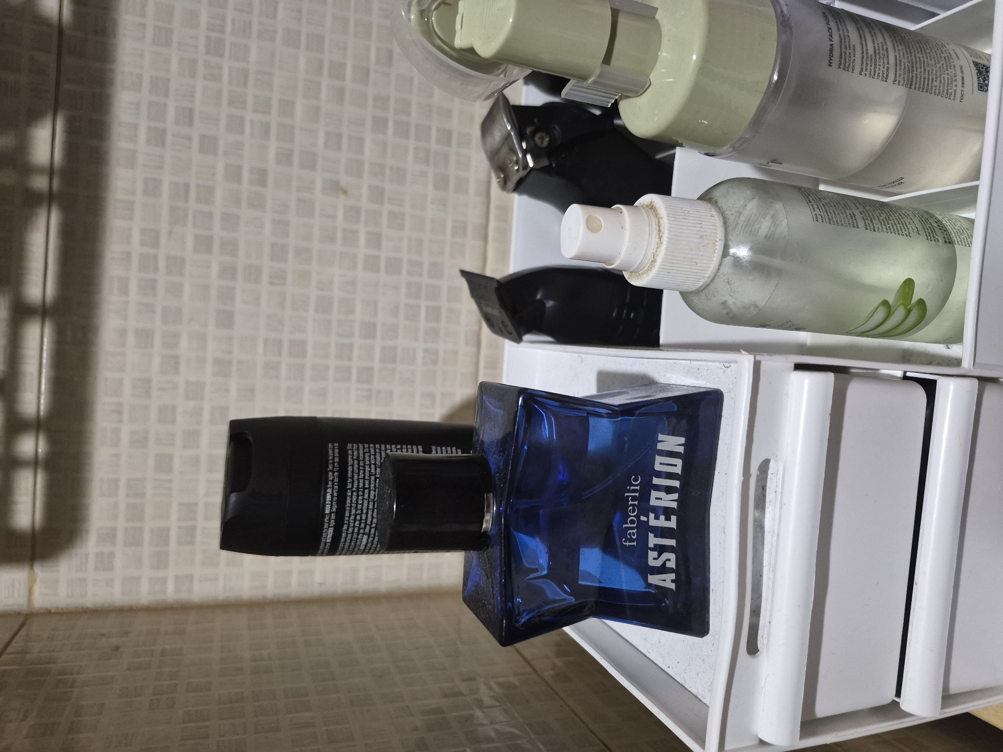 The image, rotated 90 degrees clockwise, captures a collection of personal care products organized on a white, multi-tiered shelf or organizer. The background features a light-colored tiled wall, suggesting a bathroom or dressing area.
The main subjects include several bottles and an aerosol can. Prominently displayed is a dark blue, rectangular perfume bottle with "faberlic ASTÉRION" printed in white on its side. Next to it, leaning against the perfume, is a black aerosol can. Text on this can includes phrases in English and Portuguese/French such as "for best effect," "MODE D'EMPLOI: Bien agiter...", "Segure na vertical e borrife...", "Use in well-ventilated places avoid prolonged spraying. Do not intentionally inhale..." and "Arrêter l'utilisation en cas" (stop use if).
Above these items, on a higher tier of the organizer, are two electric hair clippers or trimmers, one partially visible behind the other. To their right, a clear spray bottle with a white nozzle contains a light-colored liquid. Further up, two larger translucent bottles are visible. The lower of these appears to contain a light green liquid and features an illustration of aloe vera leaves, though most text is unreadable. The top bottle has a light green pump dispenser and visible Russian text including "HYDRA FACE FOAM," "Увлажняющая" (moisturizing), "Растительный" (vegetable/plant), "Масло жожов" (jojoba oil), and "Применение" (application).
The lighting appears artificial, casting subtle shadows on the tiled wall and within the organizer. There is no activity depicted, only a static arrangement of items. No people are visible, and no specific time of day or weather conditions can be inferred. The overall impression is a well-used but organized set of toiletries.
