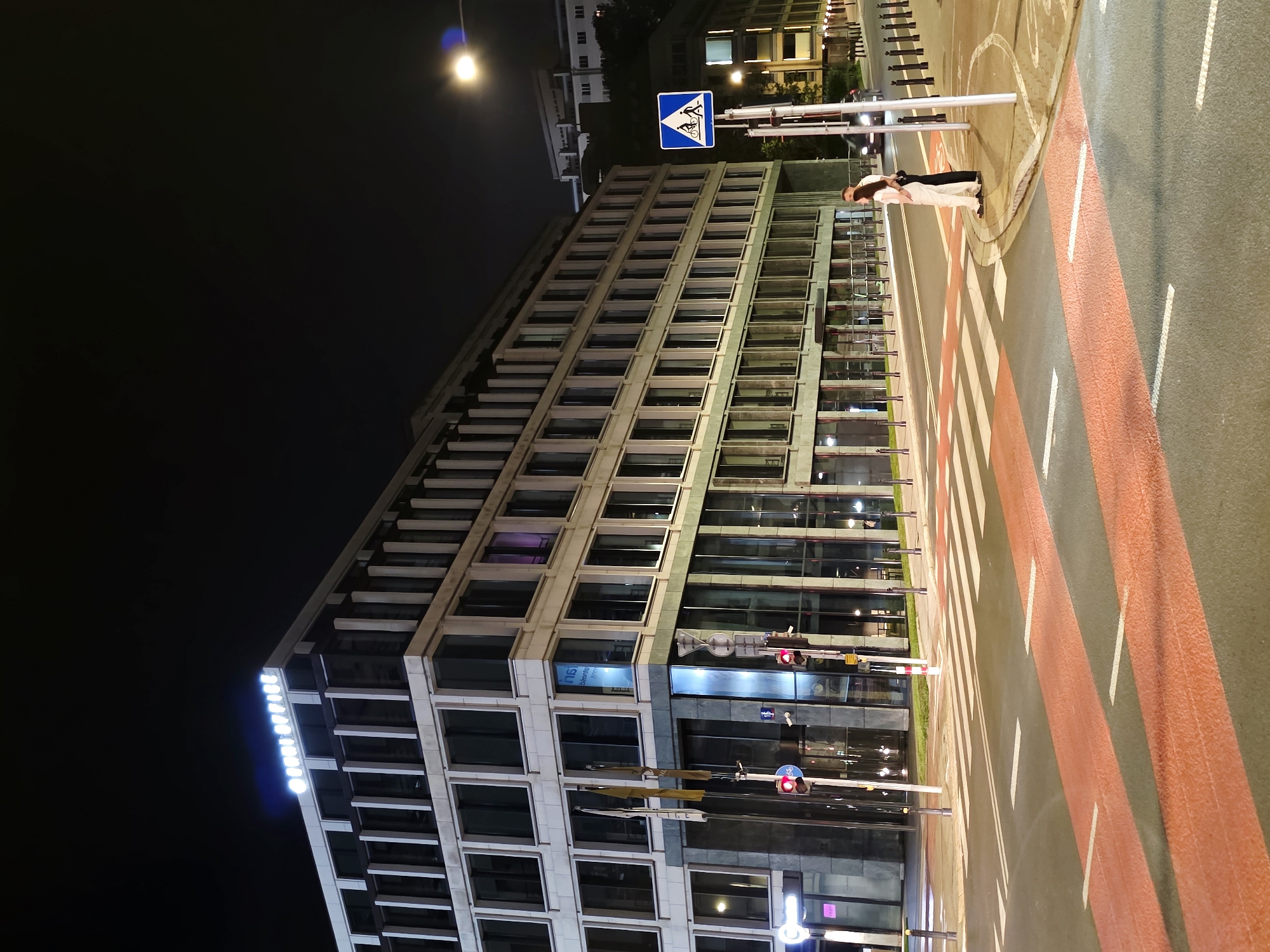 This is a nighttime, eye-level, full shot of the building with the sign "ROBI OFFICE" at the corner of Aleje Jerozolimskie and Nowy Świat streets in Warsaw, Poland. A modern, light-colored, multi-story building with a glass facade dominates the scene. The building is illuminated, with the "ROBI OFFICE" sign glowing brightly at the top. The street in front of the building has a pedestrian crossing with prominent red and white markings. Two people, a man and a woman, stand close together on the pedestrian crossing near the curb, facing the building. A streetlamp illuminates the scene, and traffic lights are visible. The sky is dark, indicating nighttime. The overall impression is one of a modern urban environment at night.