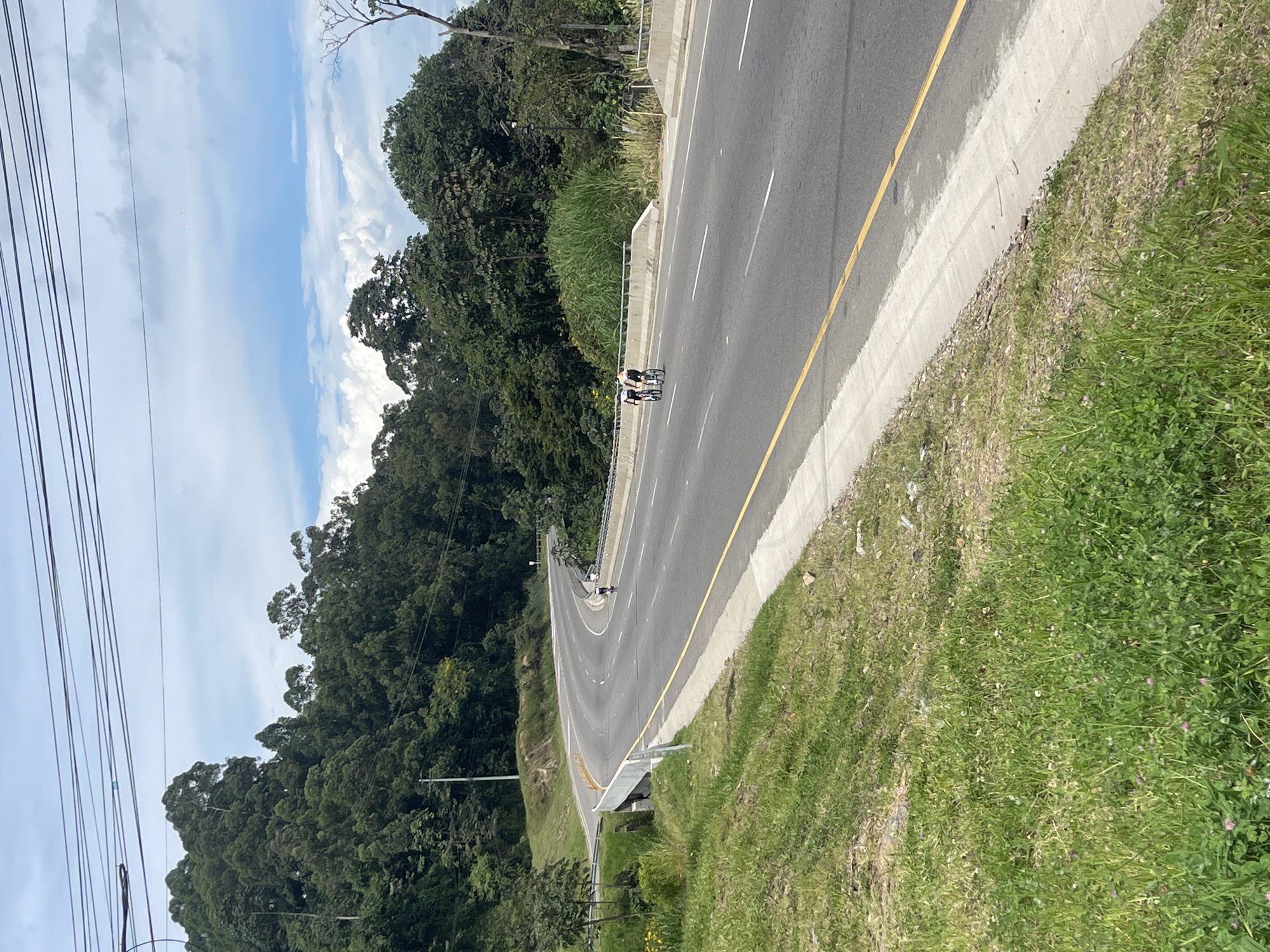 The image shows a section of highway near Silvania, Colombia. The road is asphalt, divided by solid white lines with a solid yellow line on the right edge. Concrete barriers line the shoulder on both sides of the road, which is bordered by grassy areas and dense foliage. Two cyclists are riding on the road in the distance. Utility lines are visible on the left side of the image. The sky is partially cloudy with shades of blue. The landscape is hilly and densely vegetated.