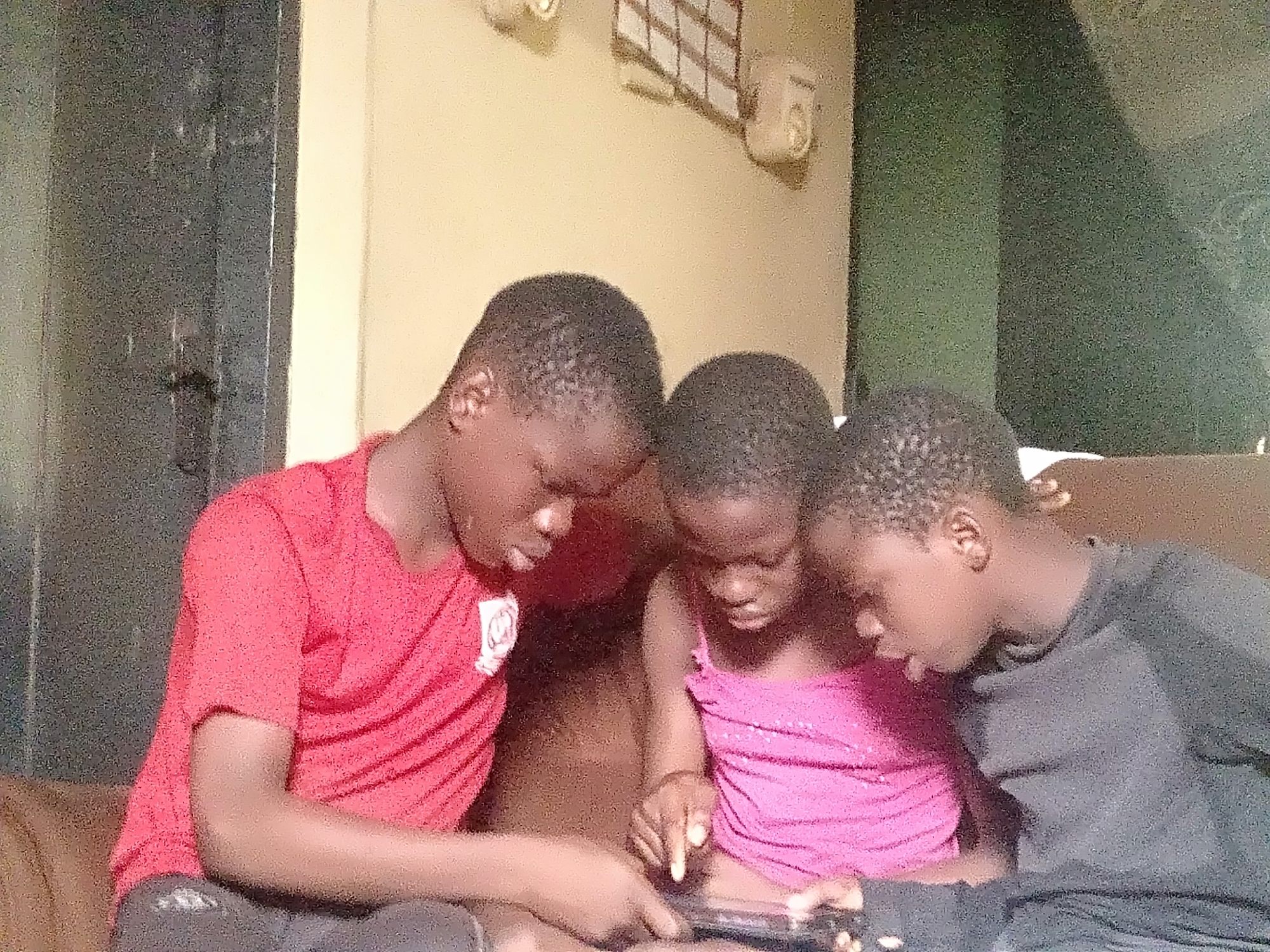 Three young children are gathered closely together on a brown sofa, engrossed in a smartphone or small tablet. The child on the left, a boy, wears a red short-sleeved t-shirt with a faint white circular logo and dark trousers. He holds the device, looking down at the screen. Next to him, a younger child, possibly a girl, in a bright pink top with sequins, actively points at the screen with her right index finger. On the right, another boy in a dark grey or black long-sleeved shirt leans in, also focused on the device. All three children appear deeply engaged and concentrated on the shared activity on the screen, suggesting they are playing a game, watching content, or interacting with an application together.
The scene takes place indoors, likely within a home in Shangisha, Nigeria. The background features light-colored, possibly tan or yellowish, walls. Visible on the wall are two light-colored wall-mounted objects, possibly fixtures or speakers, and a white grid-patterned item. A dark door or partition is partially visible on the far left, while a darker, possibly green-toned, wall section is on the right. The lighting appears dim, characteristic of an indoor setting, and the image has a somewhat grainy quality. The children's expressions show shared interest and focus. No legible text is discernible on clothing or in the background elements.