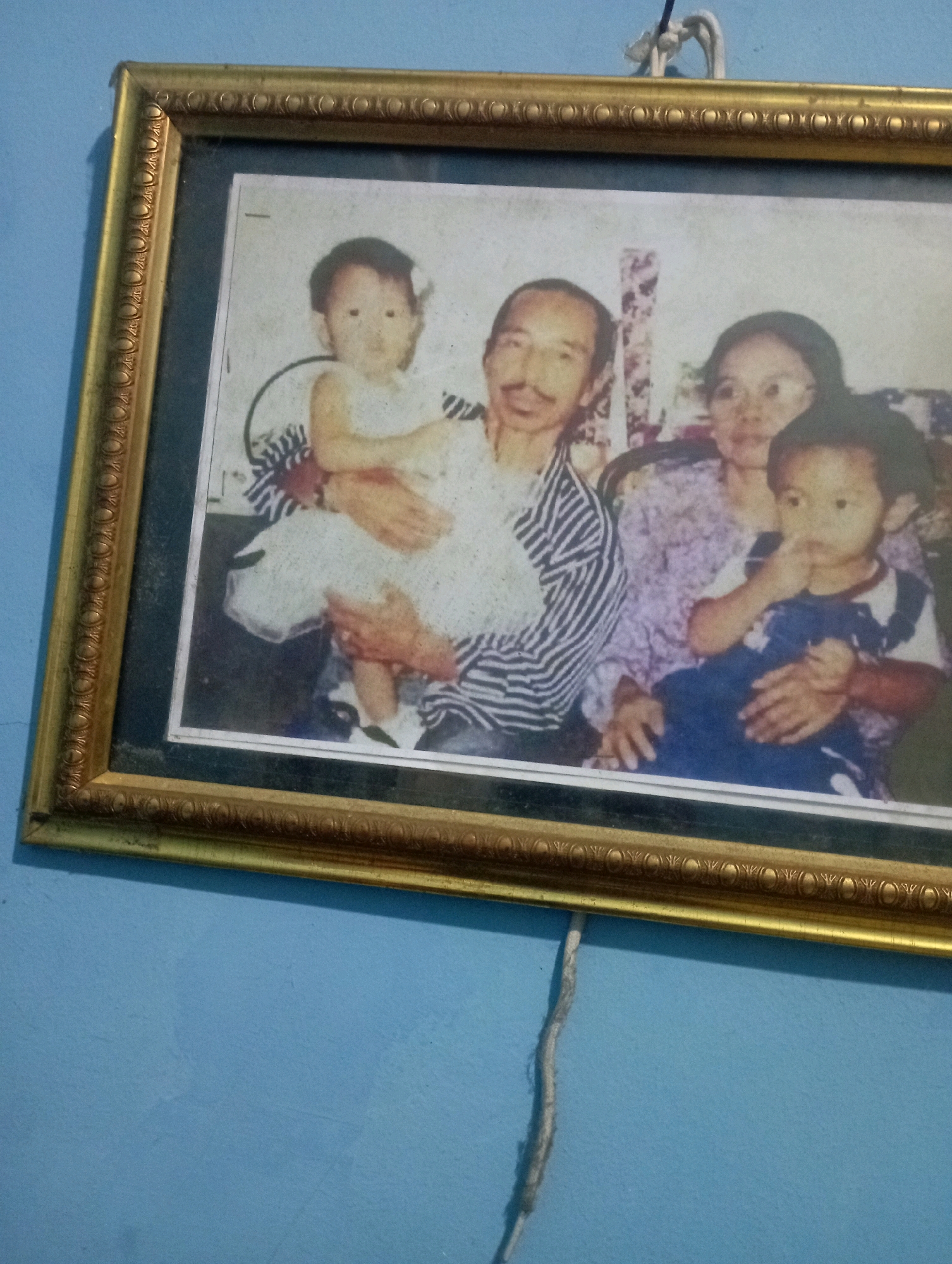 A framed photograph is displayed on a light blue wall, possibly in Senangsari, Indonesia. The photograph depicts a family of four. On the left, a man with a mustache holds a baby wearing a white dress. To the right of the man, a woman in a patterned blouse holds a second child. The background of the photograph appears to be a floral pattern. The frame is ornate and gold. A white cord hangs vertically on the wall to the left of the frame.