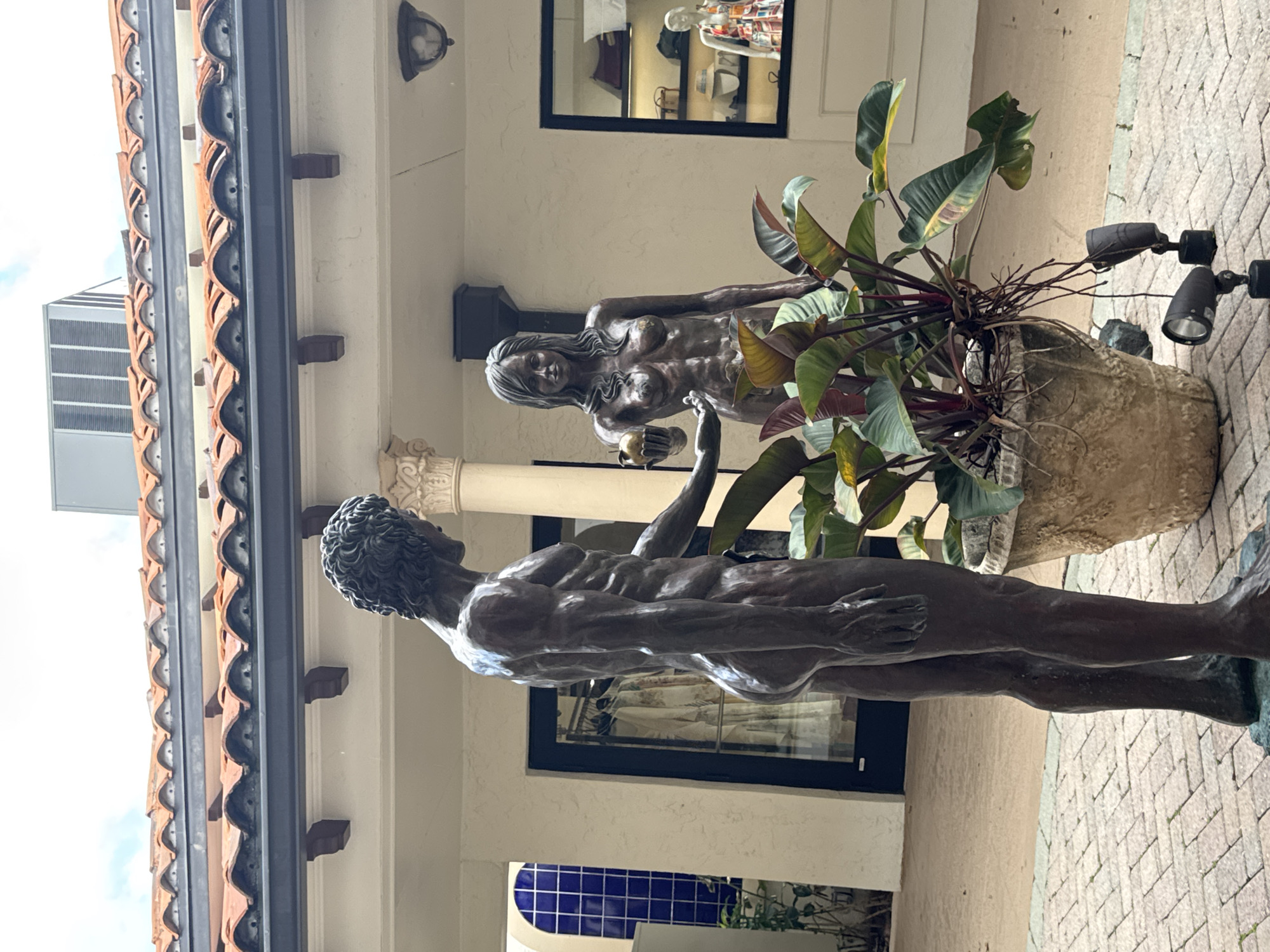This image captures an outdoor scene, likely a courtyard or plaza, featuring two bronze statues depicting a nude male and female figure, evoking Adam and Eve, in Boca Raton, United States of America.
The male statue, positioned on the left, stands facing right with his back partially towards the viewer, his right arm extended towards the female. He has a muscular physique and short, curly hair. The female statue stands to the right, facing forward, holding a spherical object, an apple, in her left hand, extending it towards the male. She possesses a slender build and long, wavy hair.
Between the two statues is a large, light-colored, textured pot containing a robust plant with large green and reddish leaves, and visible roots. Two black, ground-mounted spotlights are positioned in front of the pot, illuminating upwards. The ground is paved with rectangular light-colored brick pavers laid in a herringbone pattern.
In the background, behind the statues, is a building complex exhibiting Mediterranean Revival architecture, characterized by red Spanish barrel tile roofs and light-colored stucco walls. A dark gutter system runs below the roofline, supported by dark wooden corbels. A column with an ornate Corinthian capital is visible behind the male statue. To the far left, an archway reveals a wall tiled with small blue squares. Behind the male statue, a dark-framed glass door reflects items of clothing inside a retail space. To the right of the female statue, a dark-framed display window showcases various accessories, including hats and handbags, along with a mannequin dressed in a patterned garment. A dark downspout runs vertically on the wall behind the female statue, and a dark, dome-shaped wall light is mounted above the display window. Above the roofline on the right, a grey HVAC unit is visible against a partially cloudy sky.