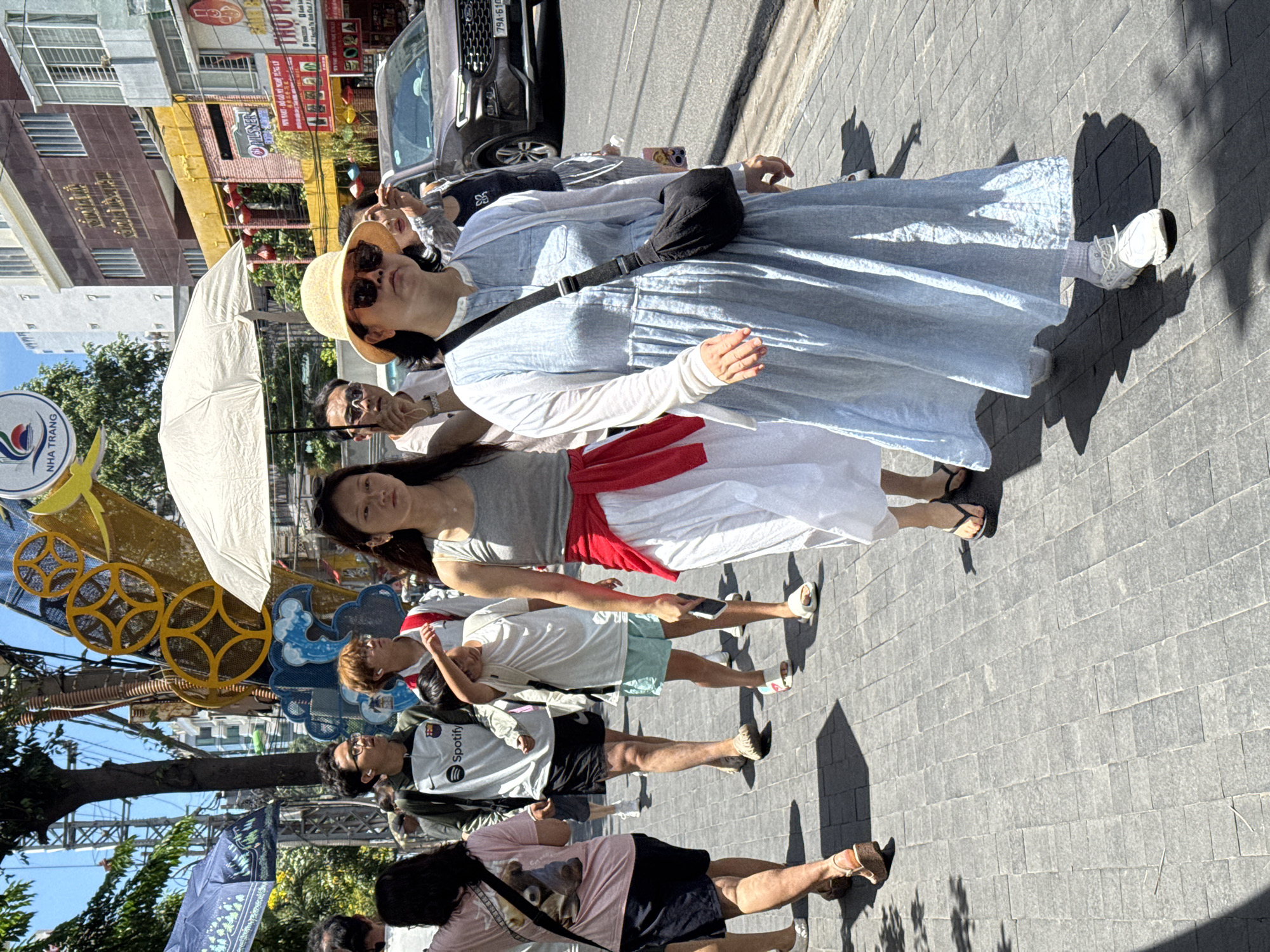 A group of people walk along a paved sidewalk in Nha Trang, Vietnam. The scene is brightly lit by the sun. In the foreground, a woman wearing a straw hat and sunglasses walks in a long, light blue dress and white sneakers. She carries a black crossbody bag. To her left, a woman in a grey tank top and white skirt, with a red sash tied around her waist, walks behind a person holding a white umbrella. Several other individuals are visible walking in the same direction, including a young person in a white t-shirt and light blue shorts, and a person in a pink t-shirt and dark shorts. Behind the pedestrians, a dark colored SUV is parked on the street, and buildings with signage are visible in the background. Decorative elements, possibly part of a festival or event, are suspended above the street, including a yellow circular design. The sidewalk is made of grey rectangular pavers.