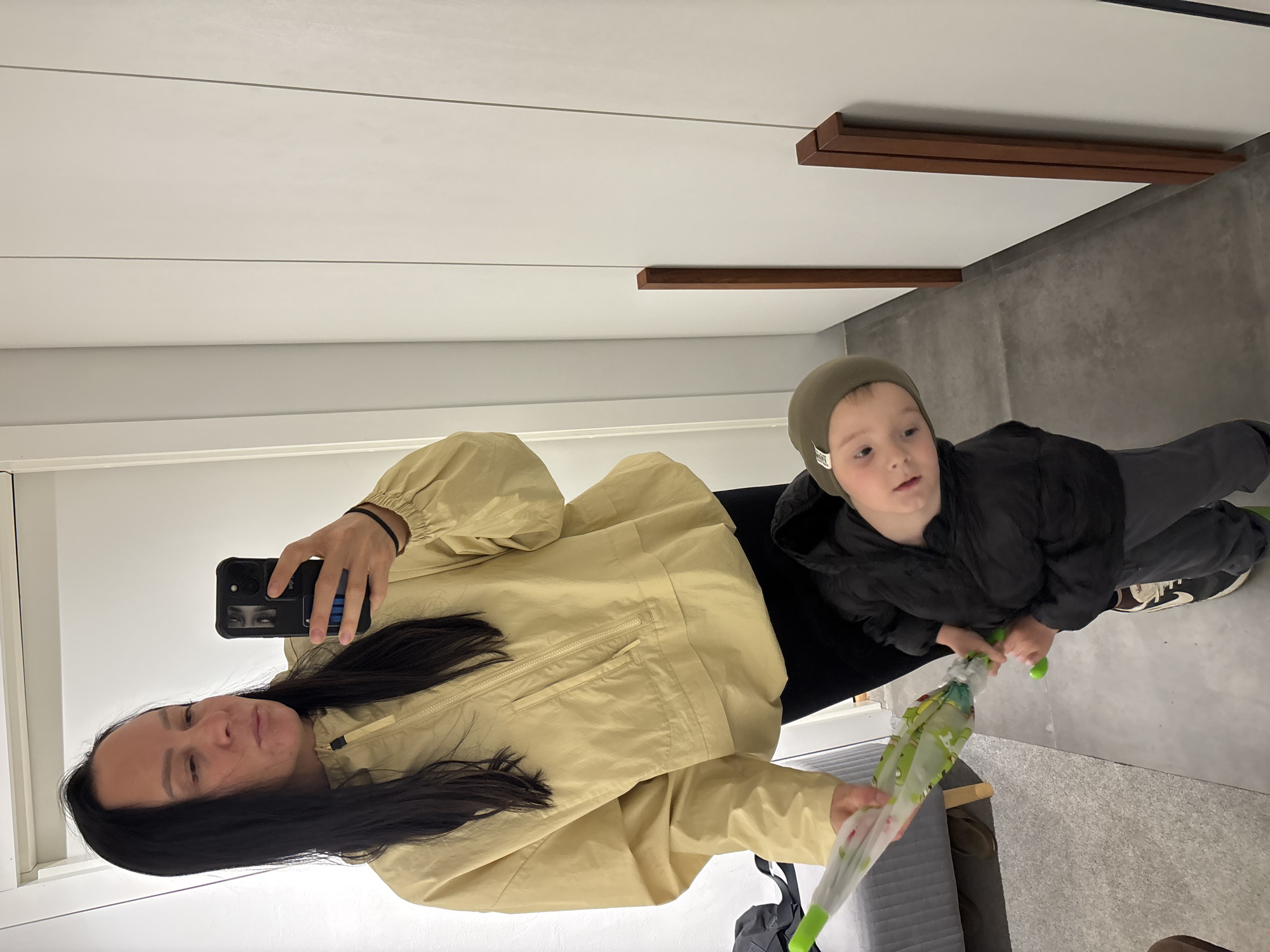 A woman and a young child are posing for a selfie in what appears to be an indoor location with modern interior design. The woman, with long dark hair, is holding up a black smartphone to take the photo. She is wearing a light yellow jacket. A reflection of her face can be seen in the phone's case. The young child, likely a boy, is standing next to her. He is wearing a dark-colored coat and a beanie-style hat. He is holding a brightly colored umbrella. The flooring is concrete or gray tile. The background features a plain white wall and a wooden beam installation. The scene is well-lit. There is nothing to indicate that the location is Chaikovsky, Russia.