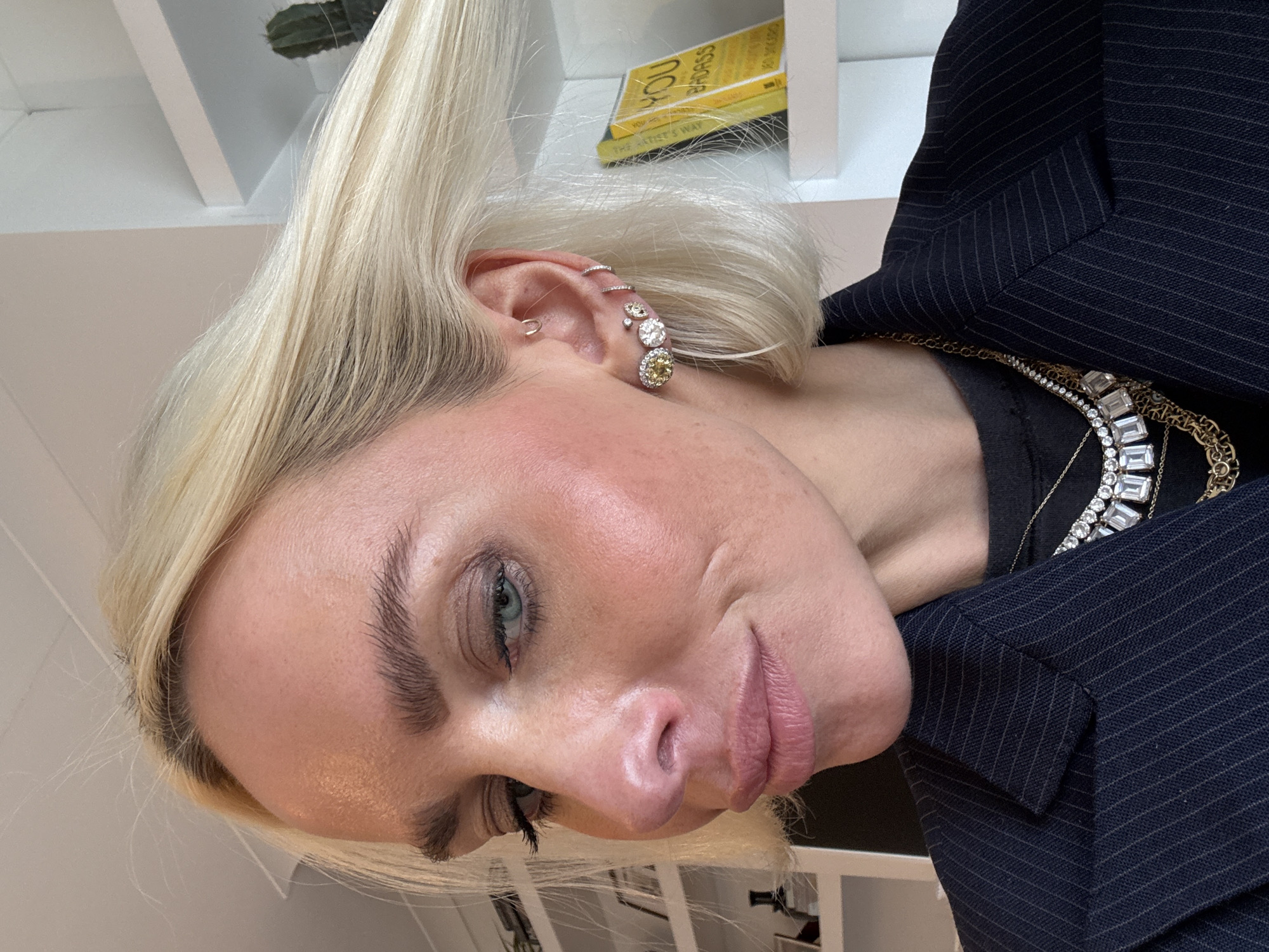 A close-up, head-and-shoulders portrait of an adult individual, likely female, with fair skin, short platinum blonde hair, and visible dark roots, is depicted. The individual's light blue-green eyes are slightly downcast, and the head is tilted. Makeup includes subtle eyeshadow, mascara, blush on the cheekbones, and a pink lip color. The individual wears a dark navy pinstriped blazer over a dark, possibly black, crewneck top. The visible right ear is adorned with multiple silver-toned earrings: an upper helix hoop, two lower helix/cartilage hoops, a small stud, and an ear climber featuring two yellow round-cut stones, one clear round-cut stone, and a smaller clear stud. Multiple necklaces are layered around the neck, including one with rectangular clear stones and another with a thicker gold-colored chain. The background is an indoor residential setting in the city of London, United Kingdom, featuring white walls and white built-in shelving. On the right, a shelf holds a small green potted cactus and a yellow book titled "YOU ARE A BADASS." On the left, additional shelves display dark-colored decorative objects, including a bottle or vase and framed items.