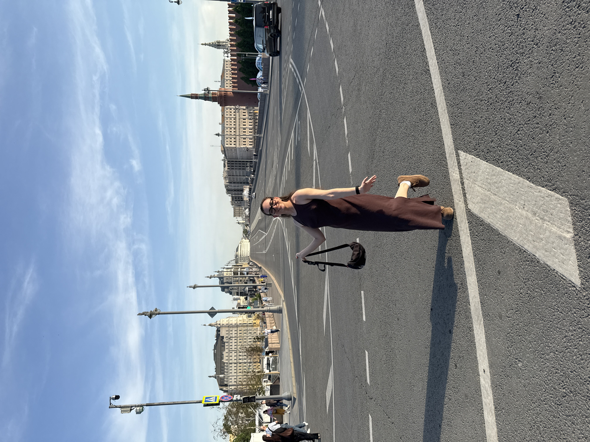 A long shot angled view of a woman standing on a roadway. The woman has on a brown dress and is wearing glasses. She's holding a purse and is midstride. The roadway has white lane markings. The background shows a city scene in Moscow, Russia. The architecture includes classic Russian towers and spires, possibly part of the Kremlin or similar structures. The sky is blue with some clouds. The time is daytime with daylight.