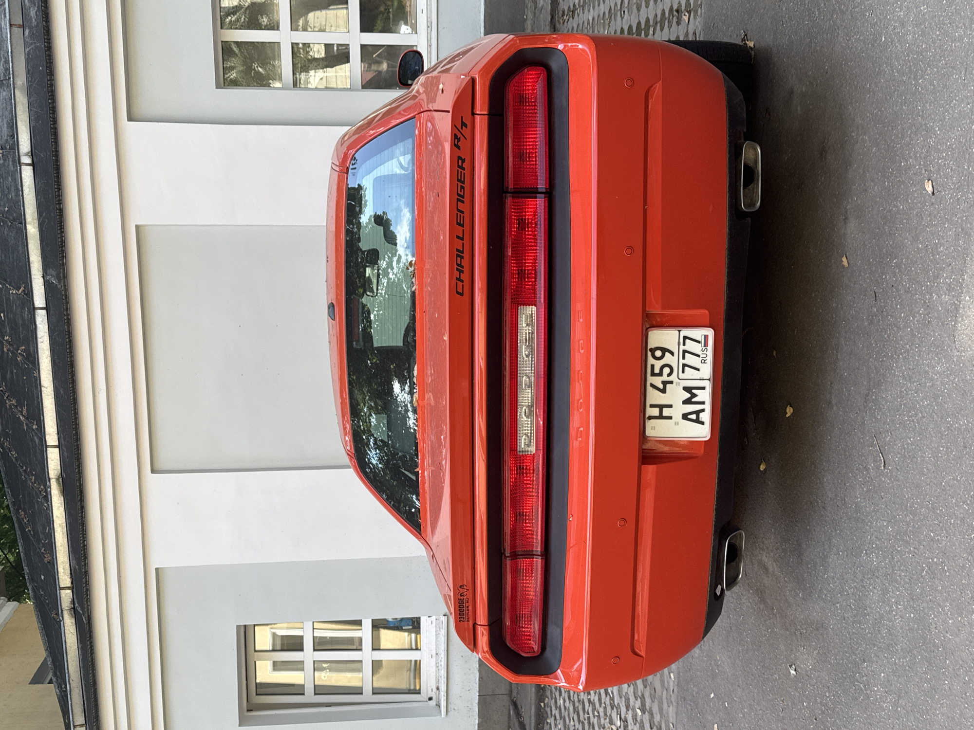 A rear view of a bright red Dodge Challenger R/T coupe is parked on a cobblestone surface in front of a white building with two windows. The car's license plate is visible, showing "H 459 AM 777" and "RUS". A small black sticker with "23DODGE BUTLER, NJ" is on the trunk, and the "CHALLENGER R/T" emblem is also present. The building has a dark roof and white decorative trim. The scene appears to be an outdoor urban environment.