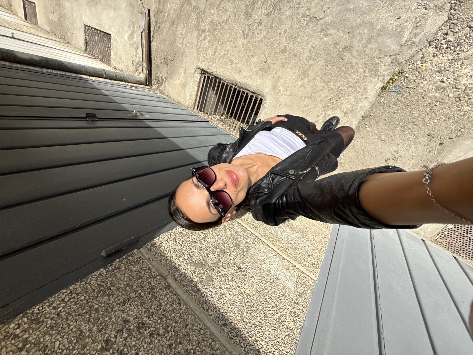 A young woman is captured in a low-angle selfie, seemingly reclining or lying on textured ground under bright sunlight in Pleven, Bulgaria. She is dressed in a black leather jacket, a white top, black shorts or a skirt, sheer black tights, and black shoes. She wears dark sunglasses, a gold chain belt around her waist, and a silver bracelet on her right wrist, which is extended to hold the camera.
The background consists of aged, textured concrete walls with two rectangular metal grates embedded within them. To the left of the woman, a dark, slatted structure, possibly a garage door or shutter, dominates part of the scene, with a metal pipe running along its top edge. The ground beneath her is a mix of small pebbles or gravel and concrete paving.
The strong, direct sunlight casts clear shadows, indicating a sunny day, likely midday or early afternoon. The setting suggests an urban, possibly utilitarian or older, outdoor environment. The woman's expression is neutral and confident. No visible text is present in the image.