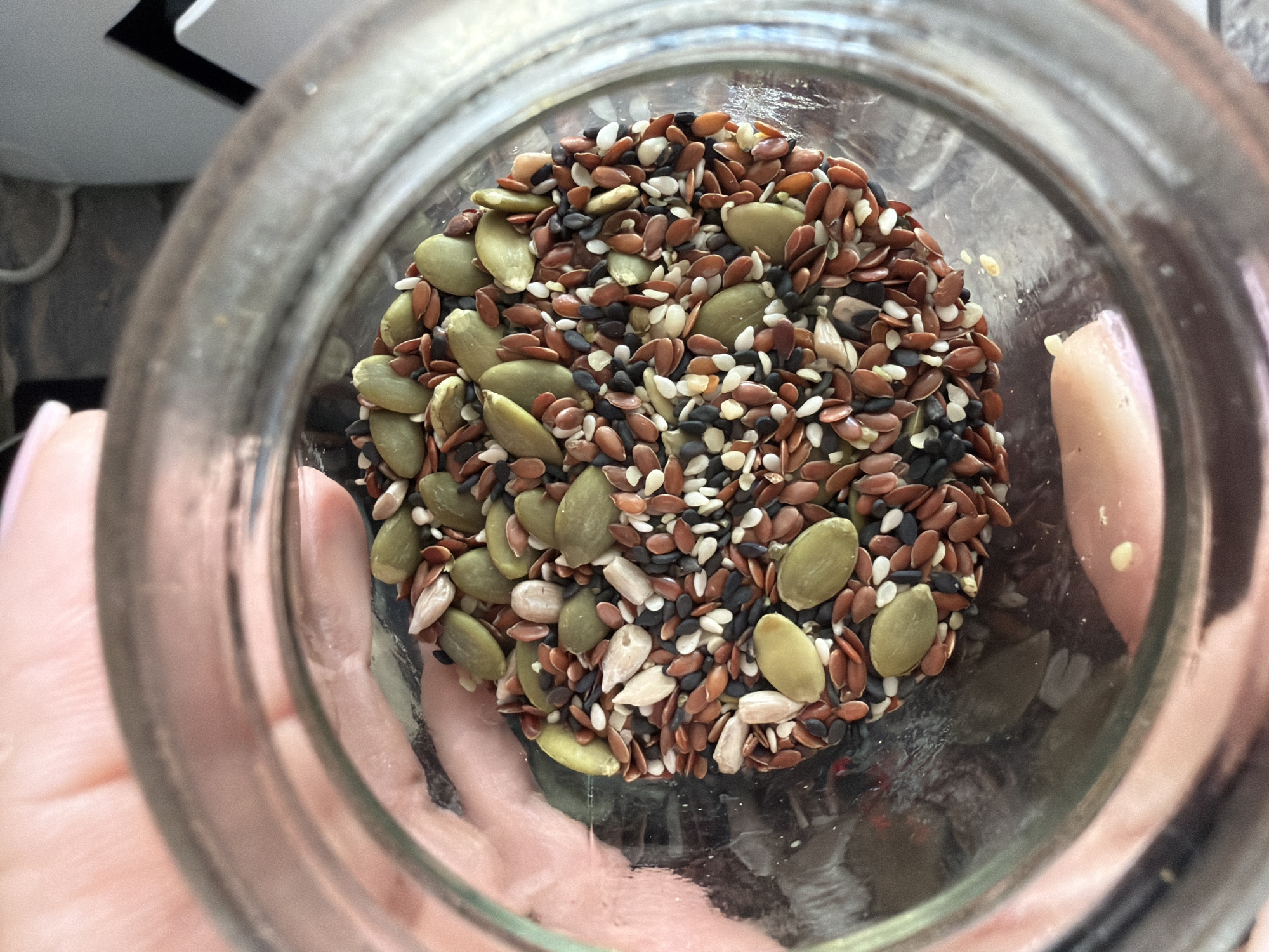 The image provides a top-down view into a clear, cylindrical glass jar, held by a person. The main subject is a diverse mixture of seeds, filling the bottom section of the jar.
The seed mix includes several types: prominent are the flat, oval, green pumpkin seeds (pepitas). Interspersed among them are numerous small, reddish-brown flaxseeds. Also visible are tiny black sesame seeds and small white or light-colored sesame seeds, contributing to a varied texture and color palette.
A person's hands, with light skin, are visible on both sides, supporting or holding the jar. The glass of the jar appears slightly smudged or dusty, particularly around the upper rim. The background, seen through and around the jar, is indistinct but suggests an indoor environment, likely a kitchen or pantry, with soft, even lighting. Hints of light-colored surfaces and dark, angular forms are vaguely discernible in the background.
The image captures a static moment, focusing on the contents of the jar. No specific activity or event is taking place beyond the observation of the seeds. There are no visible cues to indicate the time of day, weather, or any particular emotions. No text is visible within the image. While the location context is provided as Sochi, Russia, there are no visual elements in the image to confirm this.