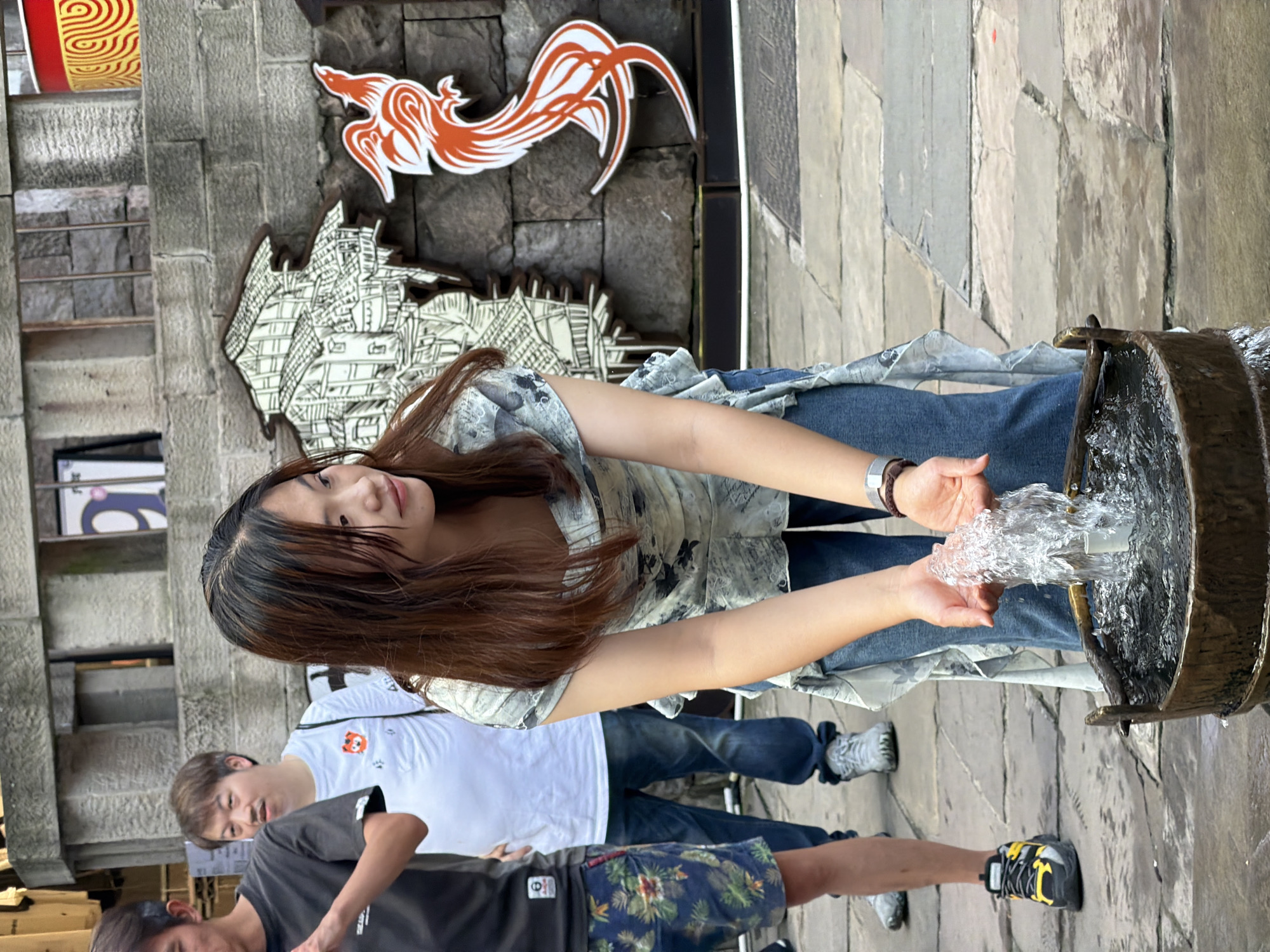 A young East Asian woman with long, dark brown hair, wearing a grey and white patterned blouse and blue jeans, is cupping water in her hands from a wooden bucket with a fountain. She is standing in what appears to be an outdoor, possibly historic, urban setting. Behind her, a man in a black t-shirt and floral shorts, and another man in a white t-shirt are visible. The background features a stone wall with decorative wooden cutouts of a building and a bird, and a red and yellow cylindrical object on a ledge. The ground is paved with stone tiles, some of which are wet. The scene suggests a public space with cultural elements, likely in Yuzhong District, China, given the context.
