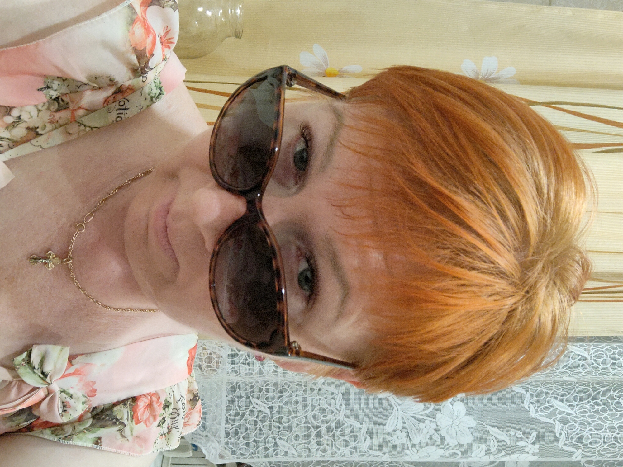 A woman with short, red hair and sunglasses is looking down at the camera, her face partially obscured by the sunglasses. She is wearing a floral-print top and a gold necklace with a cross pendant. The background consists of curtains and lace. The image was taken indoors.