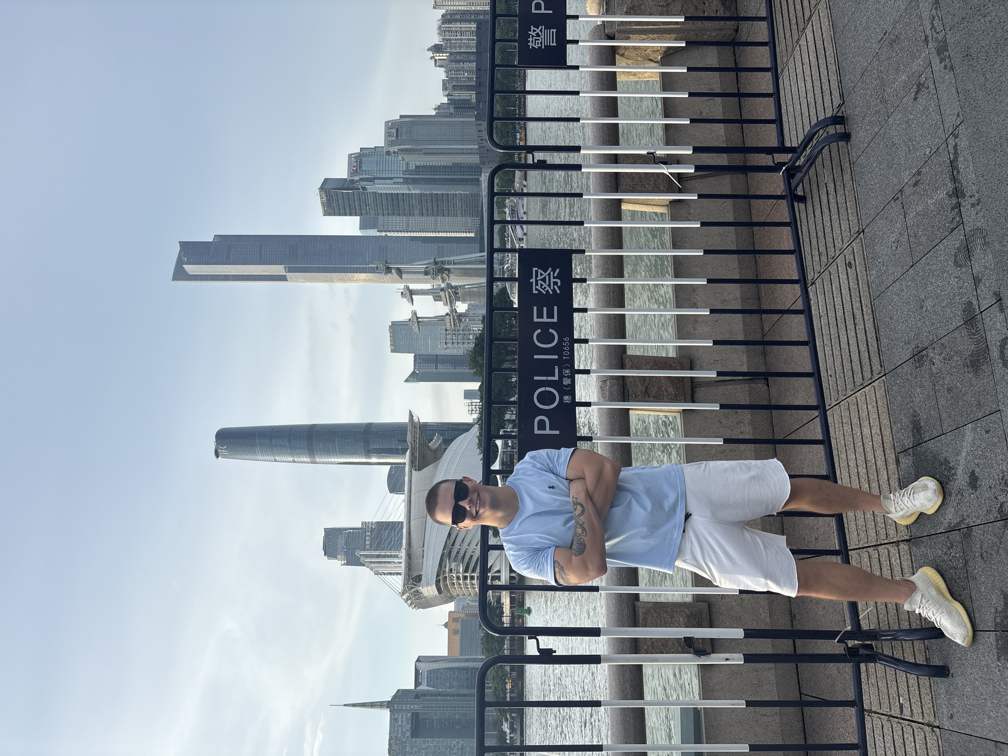 A man stands with his arms crossed in front of a police barricade on a paved walkway. The man is wearing a light blue t-shirt, white shorts, and light-colored sneakers. He has sunglasses on and his hair is short. Behind him, across a body of water, is a city skyline with numerous skyscrapers, including the Canton Tower. A boat is visible on the water. The barricade has signs that read "POLICE" in English and Chinese. The environment appears to be outdoors during the day with a clear sky. This scene is located in the Haizhu District of Guangzhou, China.