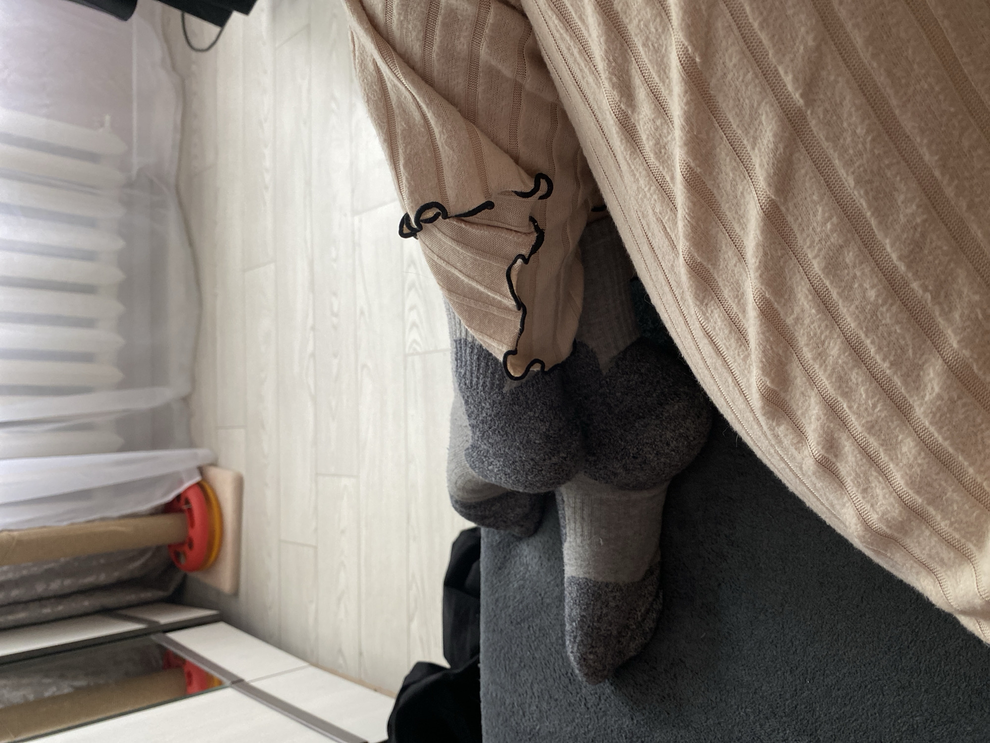 This image captures an indoor scene, likely a living room or bedroom, focusing on a person's legs and feet. The individual is wearing thick, two-toned grey socks, with darker grey on the toes and heels, and lighter grey on the main body. Their legs are partially covered by a light beige or cream-colored garment or blanket with a ribbed texture, featuring a distinctive black ruffled edge along one side. The feet are resting on a dark grey, textured surface, possibly a sofa or bed.
In the background, the floor is made of light-colored wooden planks. To the left, a window is visible, covered by white horizontal blinds and a sheer white curtain that lets in ambient light, suggesting it is daytime. Below the window, near the floor, there's a cylindrical object with red circular ends and yellow accents, positioned on a small light-colored block. Below this, reflective rectangular surfaces are visible on the floor, which could be tiles or a mirrored surface. The overall impression is one of comfort and relaxation. No text is visible in the image.
