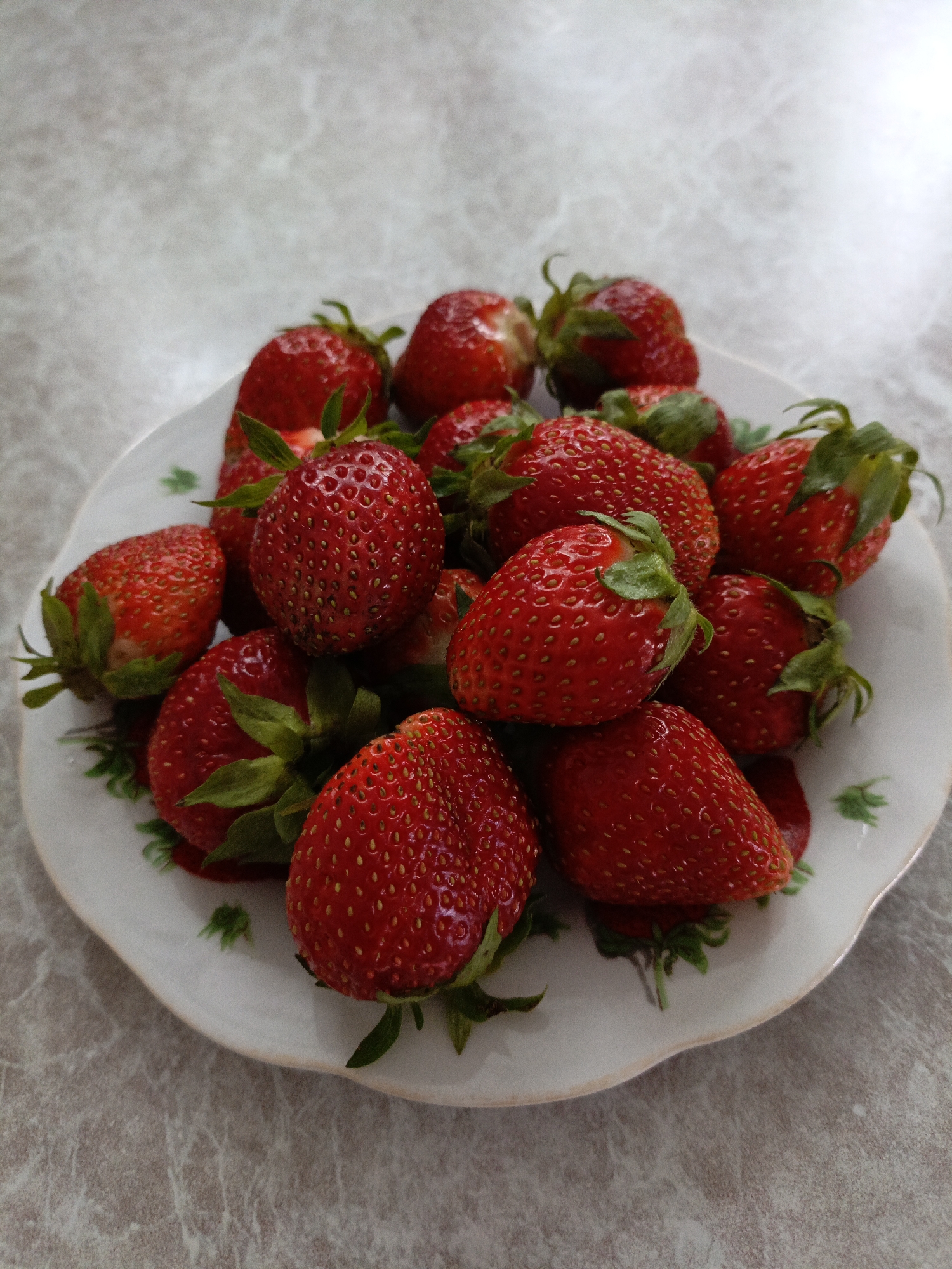 A plate of fresh strawberries is shown. The plate is white with a floral pattern and sits on a mottled gray surface. The strawberries are a vibrant red and have green stems. There are no people present in the image. The scene appears to be indoors, with the background being a blurred, neutral color. This image has no discernible connection to Kumertau, Russia.