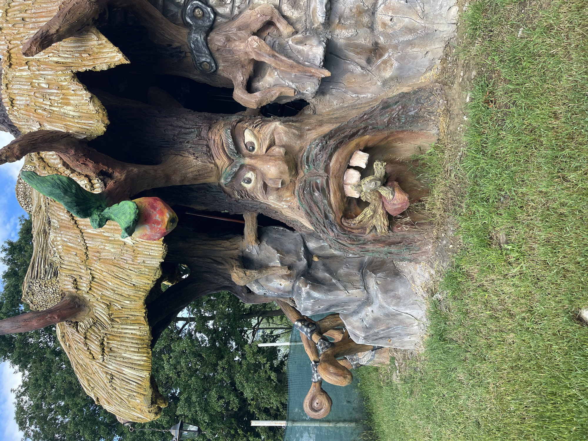 An anthropomorphic tree-like structure is prominently featured in an outdoor, grassy environment in Koprinka, Bulgaria. The structure is carved and painted to resemble multiple intertwined tree trunks with a textured, thatched-style roof in shades of yellow and brown.
The central component of the structure is a large, carved face with a grotesque expression. This face features wide, bulging eyes with dark pupils and thick, furrowed eyebrows. A large, bulbous nose is situated above a wide-open mouth, which forms an entrance into the structure. The mouth contains several large, uneven, and discolored teeth, some appearing chipped or missing. A carved mustache and beard, resembling twisted roots, frame the mouth. Inside the oral cavity, a small, green, reptilian creature with visible eyes and claws is depicted.
To the upper left of the central face, a green, organic pouch-like object, containing a red and yellow apple-like fruit, hangs from an upper branch. Further to the left, a large, stylized branch extends horizontally, adorned with two dark, metallic-banded elements and ending in a spiral shape. A dark-colored lantern hangs from the underside of the roof on the far left. To the right of the central face, another section of the structure resembles a gnarled tree trunk with a small, root-like appendage extending downwards. Above this, a dark, oval-shaped carving with two circular openings, resembling goggles or a mask, is visible. Dark recessed areas within the structure suggest interior spaces.
The structure rests on an expanse of matted green grass, which shows variations in length and some drier patches. In the background, dense green deciduous trees extend upwards towards a light blue sky with scattered white clouds. A light green mesh fence, supported by white vertical posts, is partially visible behind the structure on the left side.