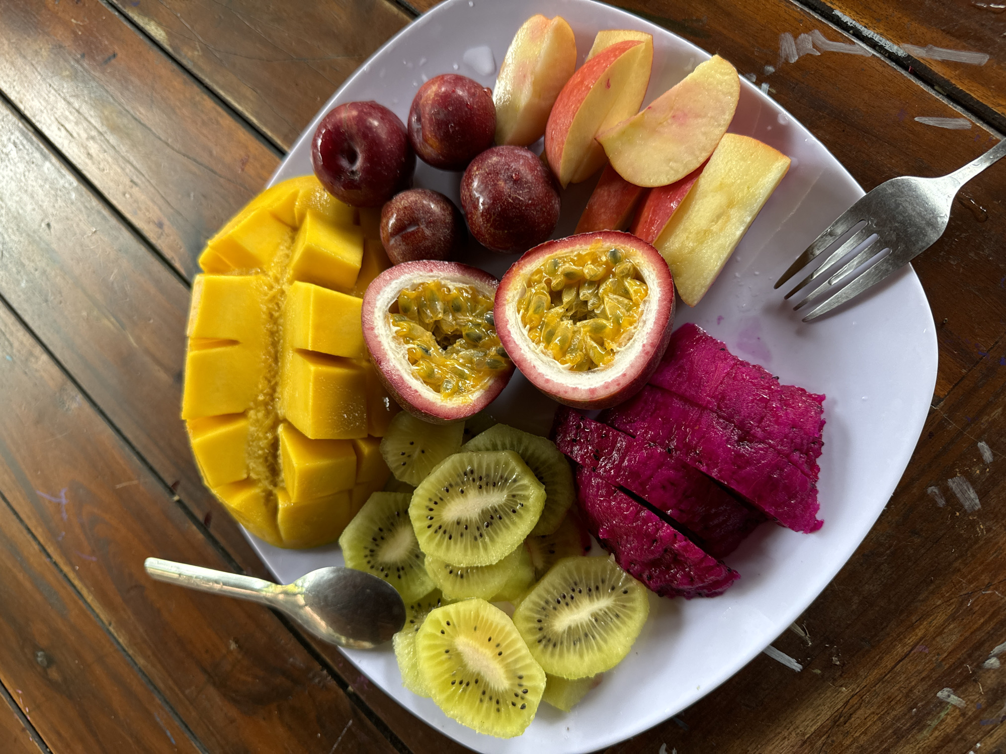 A white plate of fresh fruit rests on a weathered wooden table, likely in Mũi Né, Vietnam. The plate is arranged with: cubed mango, plums, sliced peaches, halved passion fruit with visible seeds, sliced kiwis, and wedges of dragon fruit with dark pink flesh. A metal fork and a small spoon are also on the plate. The table beneath is made of aged wooden slats with visible grain and imperfections.