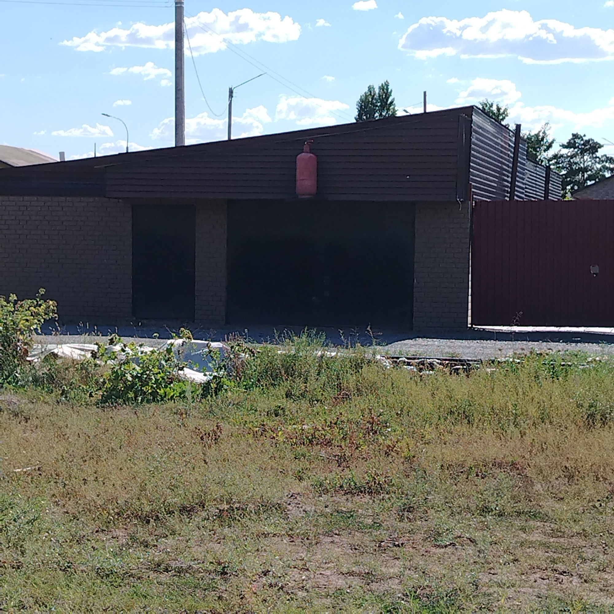The image shows a garage with two dark garage doors, constructed with brick on the sides and a dark brown roof. A red propane tank is visible on the roof. To the right of the garage is a tall, dark red metal fence. In front of the garage, there is an overgrown area with green plants and dry grass. The background consists of a blue sky with white clouds, a utility pole, and some trees. The scene is outdoors, suggesting a residential or industrial area in Ekibastuz, Kazakhstan, likely a sunny day based on the shadows.