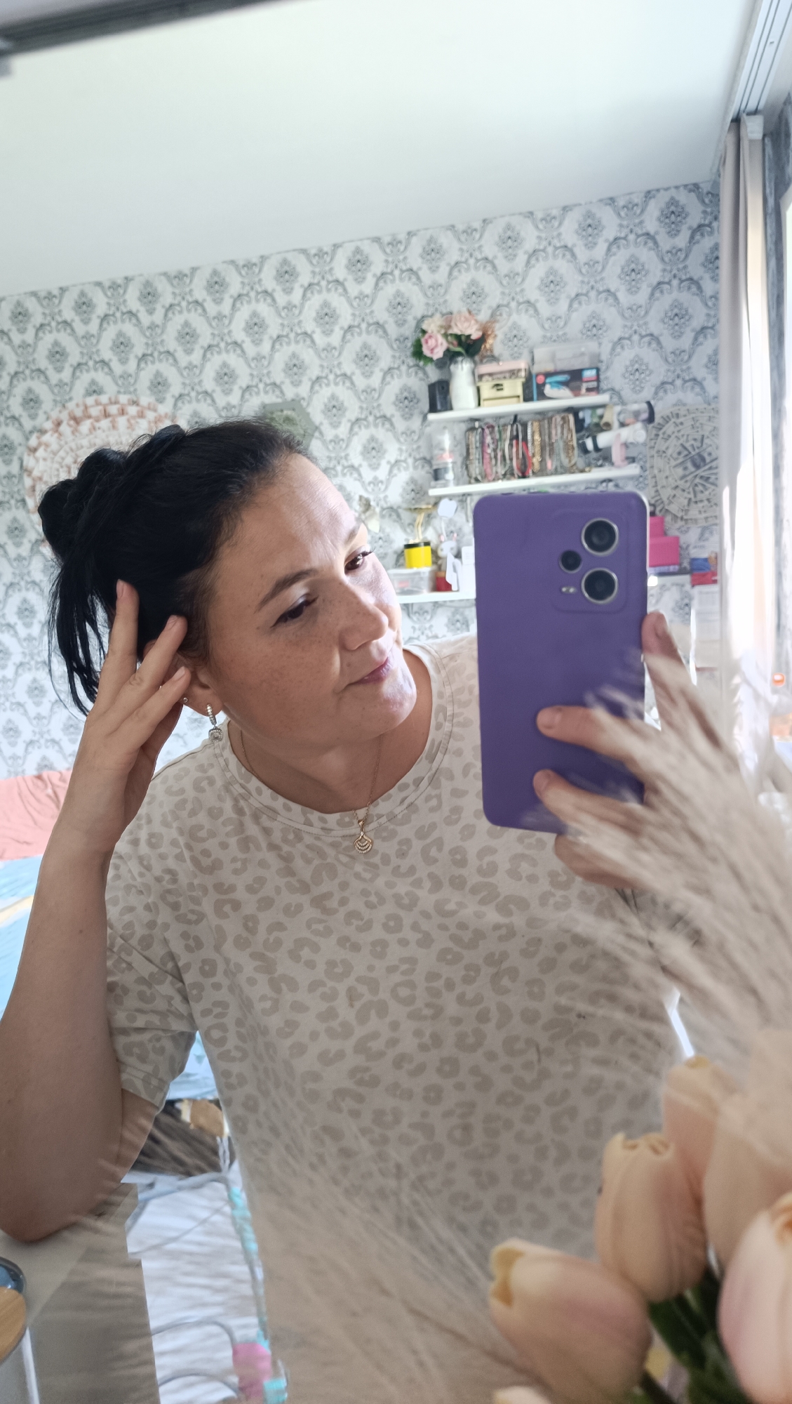A woman with dark hair pulled into a bun is taking a mirror selfie in what appears to be a bedroom. She is wearing a light-colored t-shirt with a leopard print pattern. She has her left hand on her head and is holding a purple smartphone in her right hand. Her head is slightly tilted and she is looking at her reflection in the mirror. The background shows a wall with a patterned wallpaper, shelves with various decorative items, and a window with light curtains. The room appears to be decorated in a feminine style with soft colors and floral elements. The lighting suggests it is daytime. There are no explicit indicators of the city of Chaikovsky, Russia, in the image.
