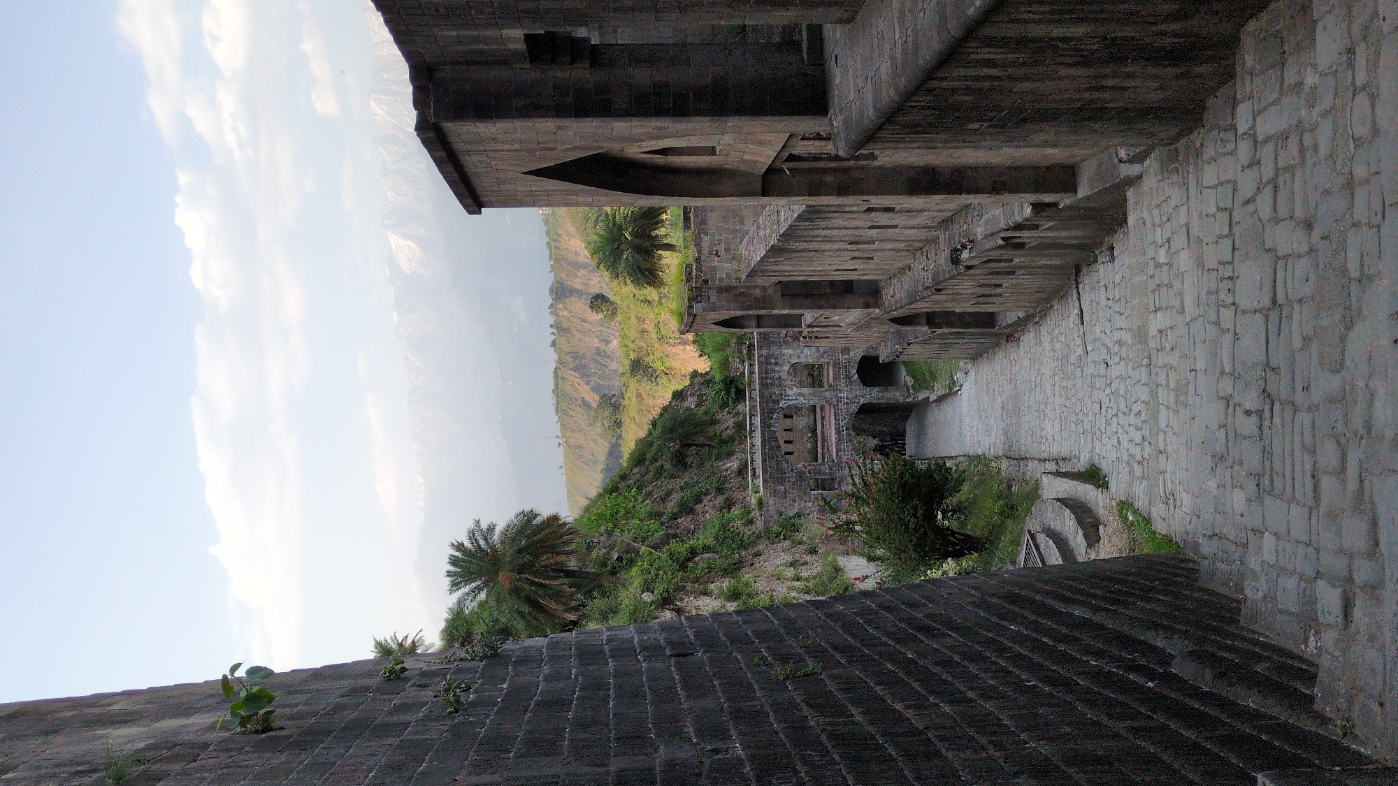 This outdoor, eye-level shot, taken in Kaswara, India, depicts a stone structure nestled near a lush, green hill. The structure, which appears to be part of an old fort or building, features a passageway of stone bricks that cuts a diagonal path through the frame. The pathway leads to a building. An upper walkway exhibits arch-shaped windows. The structure's stone is weathered and appears to be aged.
On the left side of the frame is a view of green, hilly terrain with scattered palm trees. Above the hill, the sky is visible, with some white clouds. The sky is a light blue color.
The bottom of the frame features a section of the stone structure, covered in rows of stone bricks. A few small plants can be seen growing out of the cracks in the stone.