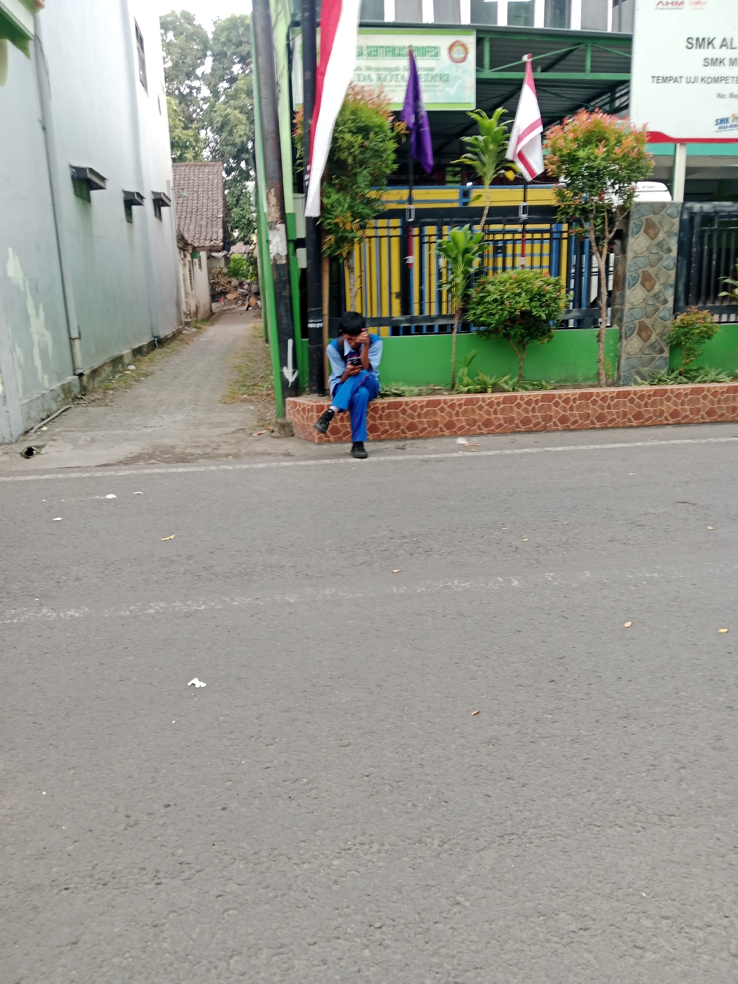 A young male wearing a blue school uniform with white accents sits on a textured, reddish-brown stone bench on the side of a paved road. He is looking down at a mobile phone in his hands. The background shows a building with green and yellow striped fencing, flags, and trees. A narrow alleyway is visible to the left of the building, with a white wall of another building adjacent to it. The scene appears to be in an urban or suburban setting in Kediri City, Indonesia, likely near a school given the student's attire. The road surface is asphalt, and scattered leaves are visible on the road.