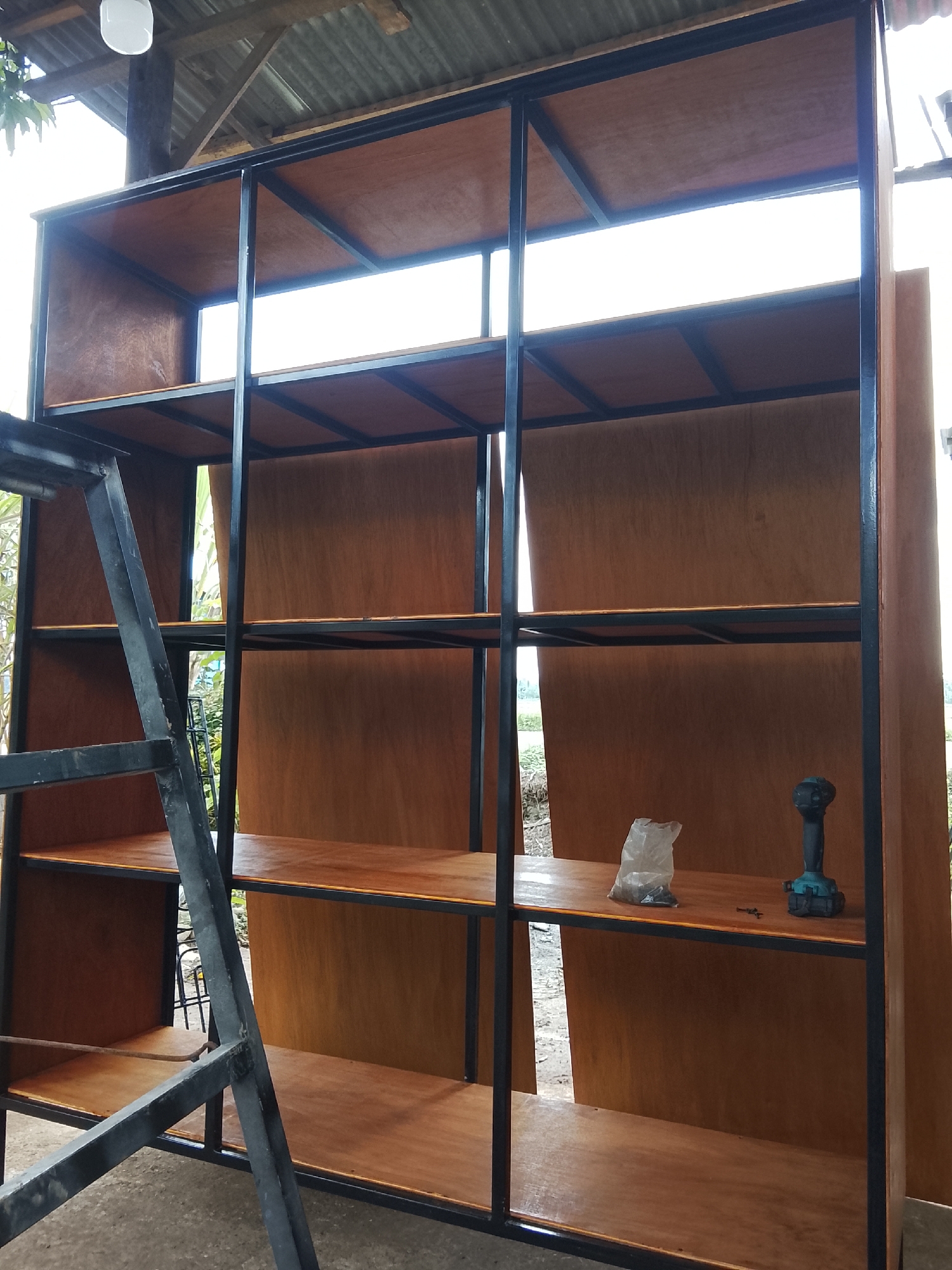A large, unfinished wooden and metal shelving unit is being assembled outdoors, under a corrugated metal roof supported by wooden beams. The unit has multiple shelves made of polished plywood, framed by black metal supports. A metal ladder leans against the left side of the shelving unit. On the second shelf from the top, a clear plastic bag filled with small items, a few screws, and a teal-colored power drill are visible. The background shows greenery and a distant horizon. The scene is likely in Mee, Indonesia, given the architectural style of the roof and the surrounding vegetation.