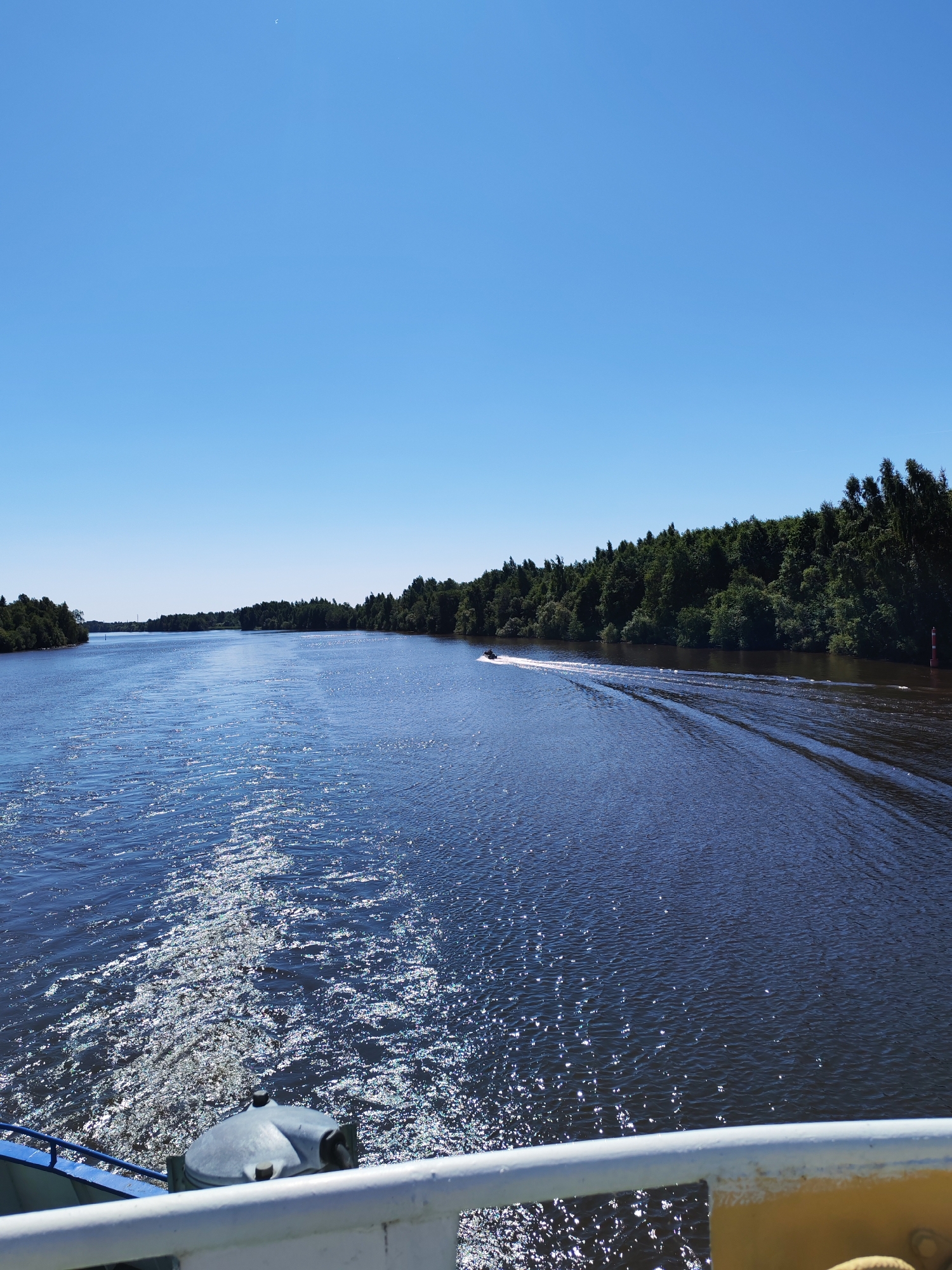 The image depicts a wide, dark blue river under a clear, bright blue sky. Lush green trees line the banks of the river on both sides, extending into the distance. A jet ski is visible in the middle ground, leaving a white wake on the water. The foreground shows a portion of a boat, with a white railing and a grey capstan or winch. The water sparkles with sunlight, indicating a sunny day. The scene appears to be a tranquil, natural environment, likely in or around Vytegra, Russia, given the context of the city being a port on the Vytegra River and part of the Volga-Baltic Waterway.