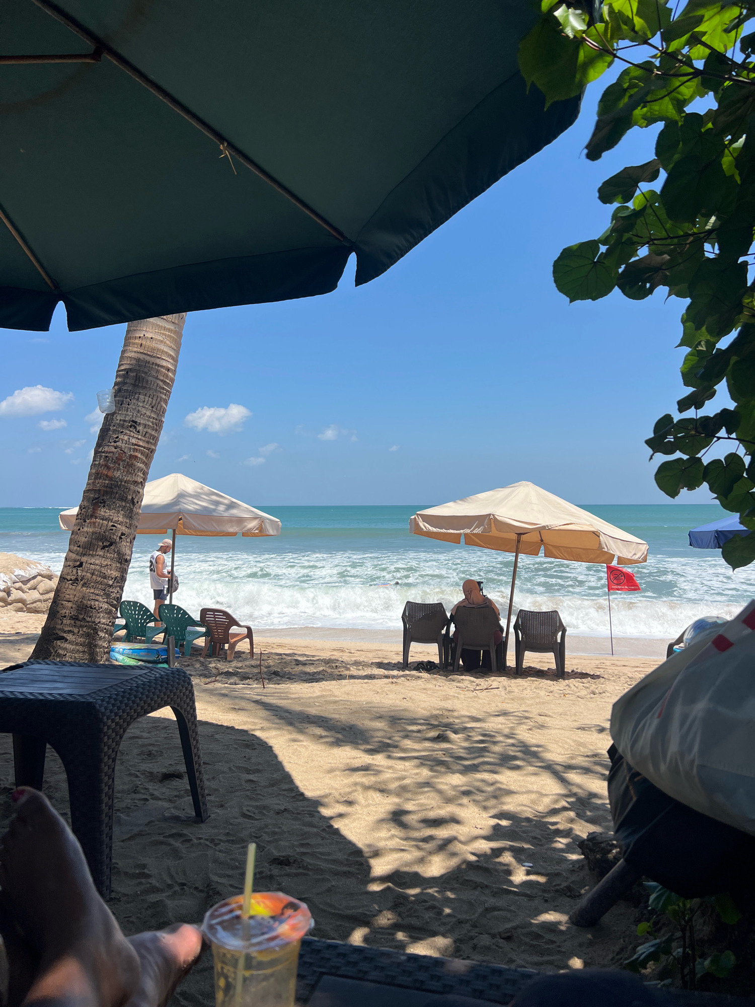 The image depicts a beach scene in Kuta, Indonesia. In the foreground, a person's legs are visible, resting on a dark-colored chair, with a partially consumed beverage in a clear plastic cup with a yellow straw situated on a table. The background reveals a sandy beach with several beach chairs and umbrellas. Two beach umbrellas, one large beige and another smaller white one, are set up. Under the beige umbrella, a person wearing a headscarf and light-colored clothing is seated, facing the ocean. To the left, near the white umbrella and a palm tree, another person in a white tank top and hat stands looking at their phone. The ocean waves are active, with white foam indicating movement. A red flag is planted on the beach near the beige umbrella. Trees with green leaves are visible in the upper right corner, casting shadows on the sand, along with the dark green canopy of another umbrella in the upper left. The sky is clear blue with scattered white clouds.