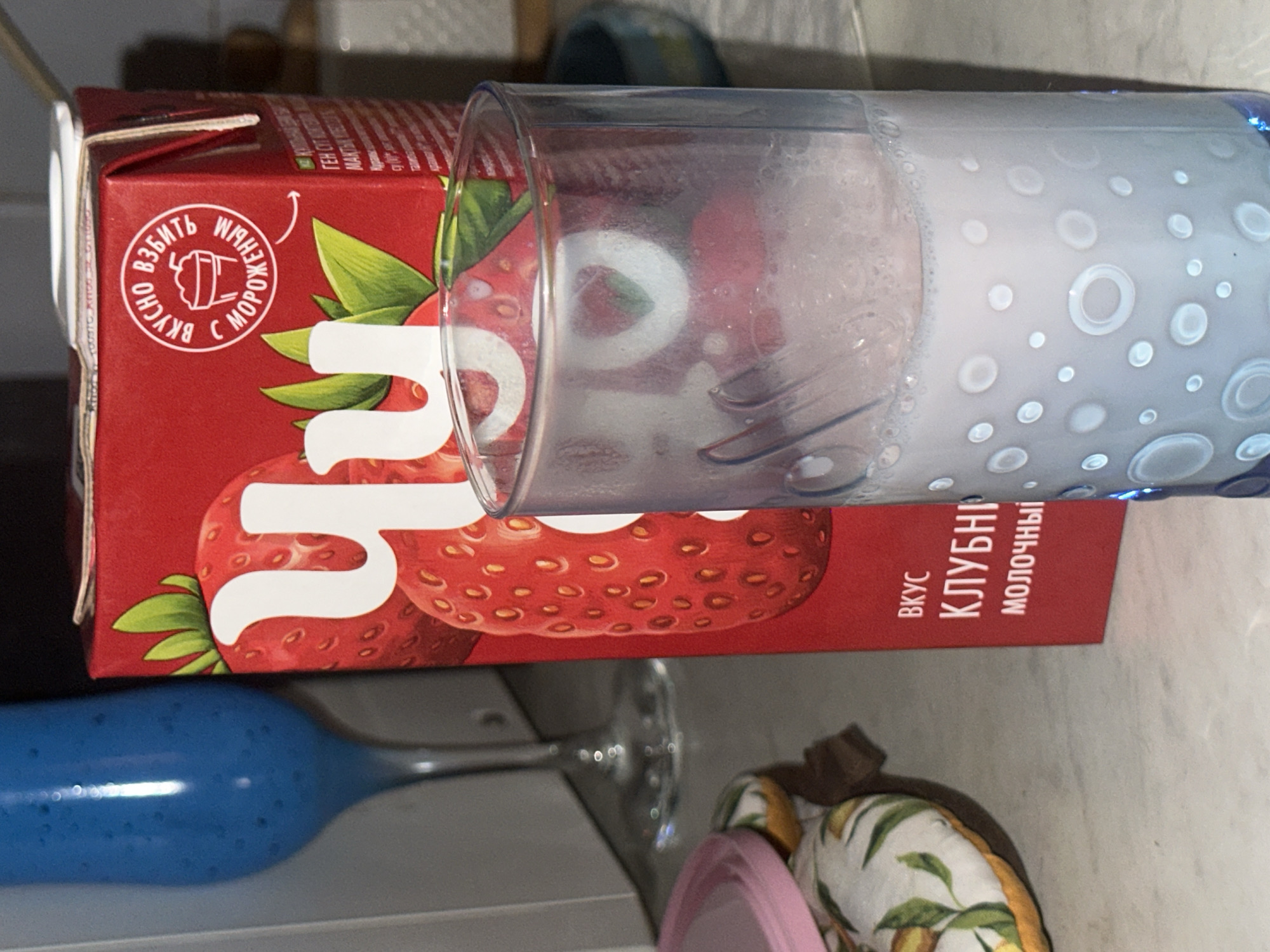 The image displays an indoor scene, likely a kitchen, situated in the city of Moscow, Russia, as indicated by the Russian language text on the product packaging.
In the center-right, a tall, red cardboard carton stands upright. The carton features a large graphic of strawberries and Russian text. A white circular emblem on the upper portion reads "ВКУСНО ВЗБИТЬ С МОРОЖЕНЫМ" (TASTY TO WHIP WITH ICE CREAM). Below this, at the bottom-front, additional text states "ВКУС КЛУБНИ" (STRAWBERRY FLAVOR) and "МОЛОЧНЫЙ" (DAIRY/MILK). A large Cyrillic letter "Ч" (Ch), characteristic of the "Чудо" brand, is prominently displayed on the strawberry graphic.
Immediately in front of the carton, a clear cylindrical glass contains a light-colored, frothy liquid. The glass is adorned with a pattern of translucent circular and ring-shaped indentations. A blue stemmed glass, filled with a dark blue or purplish liquid, stands in the background on the left side.
To the bottom-left, a fabric item, possibly a potholder or oven mitt, is visible. It features a pattern of yellow lemons and green leaves on a light background. A pink, circular lid rests partially on this fabric item. In the far right background, a small, dark blue or green bowl is partially visible.
The objects rest on a light-colored countertop. The background consists of a plain, lighter-toned wall or cabinet. No individuals are present in the scene. The arrangement of the items suggests the preparation or consumption of beverages, specifically milkshakes.