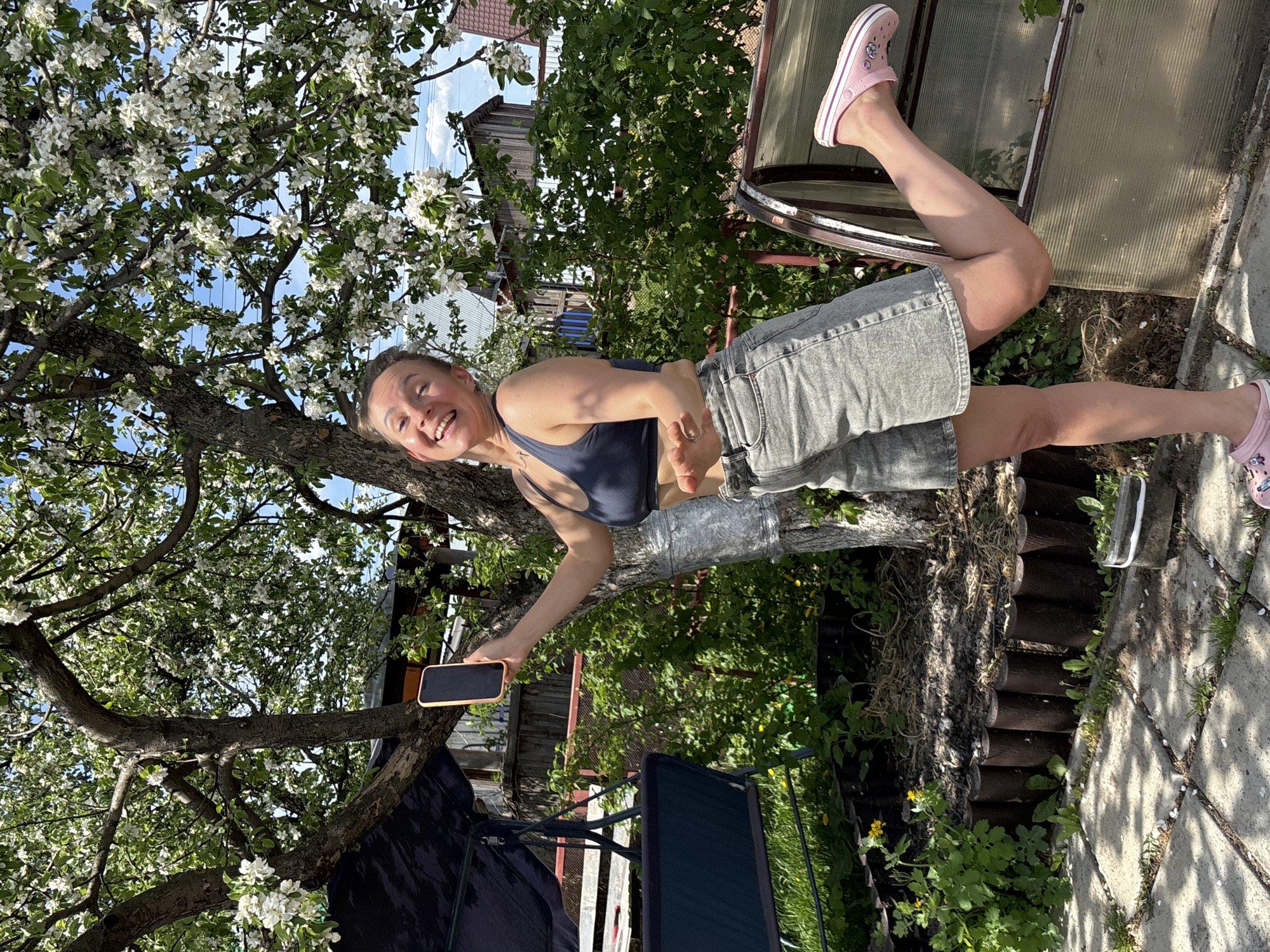 A woman smiles and holds a smartphone while standing on the trunk of a flowering tree in what appears to be a yard in Chaikovsky, Russia. She wears a dark sports bra, denim shorts, and pink clogs. The yard includes a greenhouse-like structure, a wooden fence, garden furniture, and vegetation. The tree's white blossoms are prominent against a partly cloudy sky.