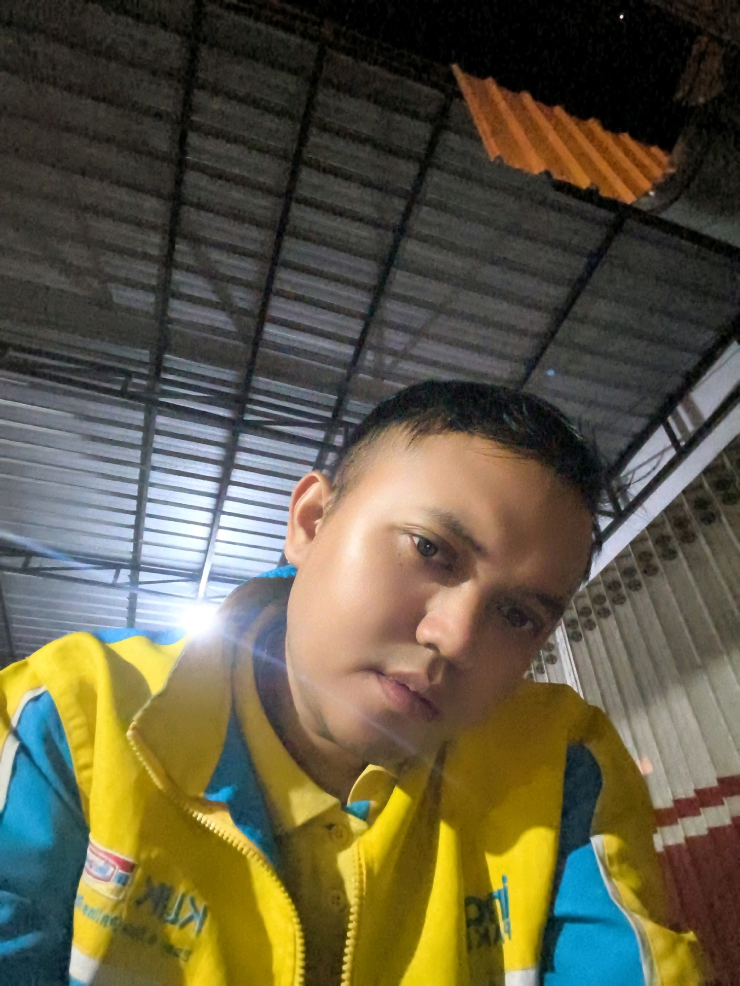 A young East Asian male is captured in a low-angle, close-up selfie, wearing a bright yellow jacket with blue accents over a yellow collared shirt. The jacket features a zipper and embroidered logos. The man has short dark hair and is looking directly at the camera with a neutral expression. The background consists of a metal roof structure with visible beams and corrugated metal sheeting. To the right, there appears to be a wall with a repeating pattern of white and red stripes. The lighting suggests it is nighttime or a dimly lit indoor space. The location is identified as Karangtalun, Indonesia.
