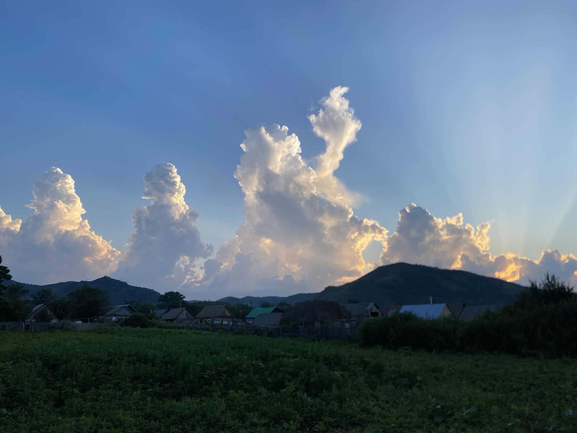The image depicts a rural landscape at dusk, likely in Maloarslangulovo, Russia. The sky is a vibrant blue, with dramatic, illuminated clouds that appear to be shaped like figures. Sunlight streams through these clouds, casting visible rays across the scene. Below the sky, a range of dark, silhouetted mountains forms the horizon. In the mid-ground, a cluster of small, traditional houses with wooden fences are visible, surrounded by trees and a large haystack. The foreground is dominated by a dense field of dark green vegetation, possibly crops or a meadow, which occupies the lower half of the frame. The overall impression is one of natural beauty and rural tranquility at the end of the day.
