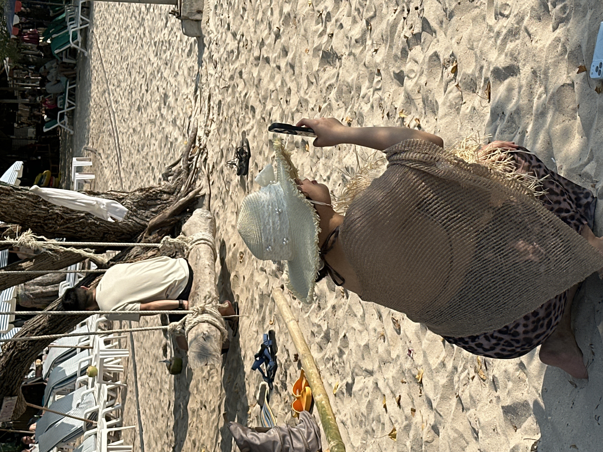 A sunny, sandy beach scene in Pattaya City, Thailand, presents an older woman kneeling on the sand, taking a picture with a smartphone. She wears a straw sunhat, a brown mesh top, and leopard-print shorts. To the left, a man in a light-colored shirt and dark shorts lies prone, resting on a structure of bound logs with ropes. Stacked white plastic chairs fill the background, and beachgoers lounge on green and white chairs in the distance. The beach sand shows scattered leaves. Personal items, including footwear and clothing, are strewn around the log structure.