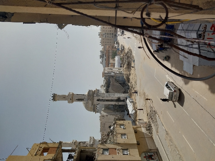 The image depicts a street scene in Khan Yunis, Palestinian Territory, characterized by extensive urban damage under a clear daytime sky. A prominent mosque with a light-colored minaret and dome, partially damaged with sections missing and debris at its base, anchors the center background. To the left, a multi-story building is severely compromised, with its facade and internal structures collapsed, exposing rubble. Another damaged building with exposed rebar and hanging wires frames the right foreground.
The street is an asphalt road with faded white markings. A silver-colored station wagon is either parked or moving slowly in the right lane, facing away from the viewer. Approximately 10-15 individuals are present, walking on the street and sidewalks; one person walks towards the vehicle, and another pushes a cart near temporary structures on the right side of the road, which display Arabic script. Large piles of concrete and building debris are scattered along the street's edges and near the damaged structures. Overhead, multiple thin wires stretch across the street.